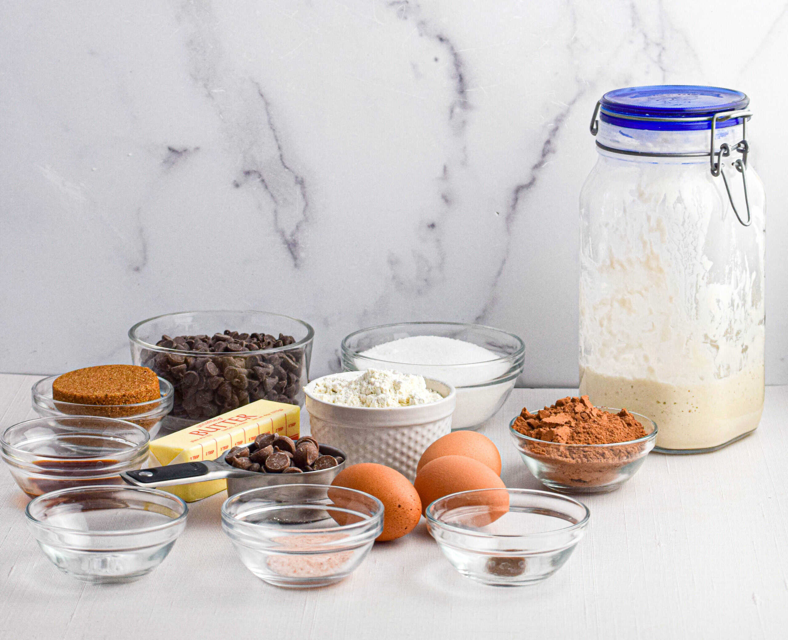 Ingredients for sourdough brownies are laid out on a white backdrop, including a jar of sourdough starter, eggs, cocoa powder, flour, sugar, chocolate chips, vanilla extract, butter and salt. 