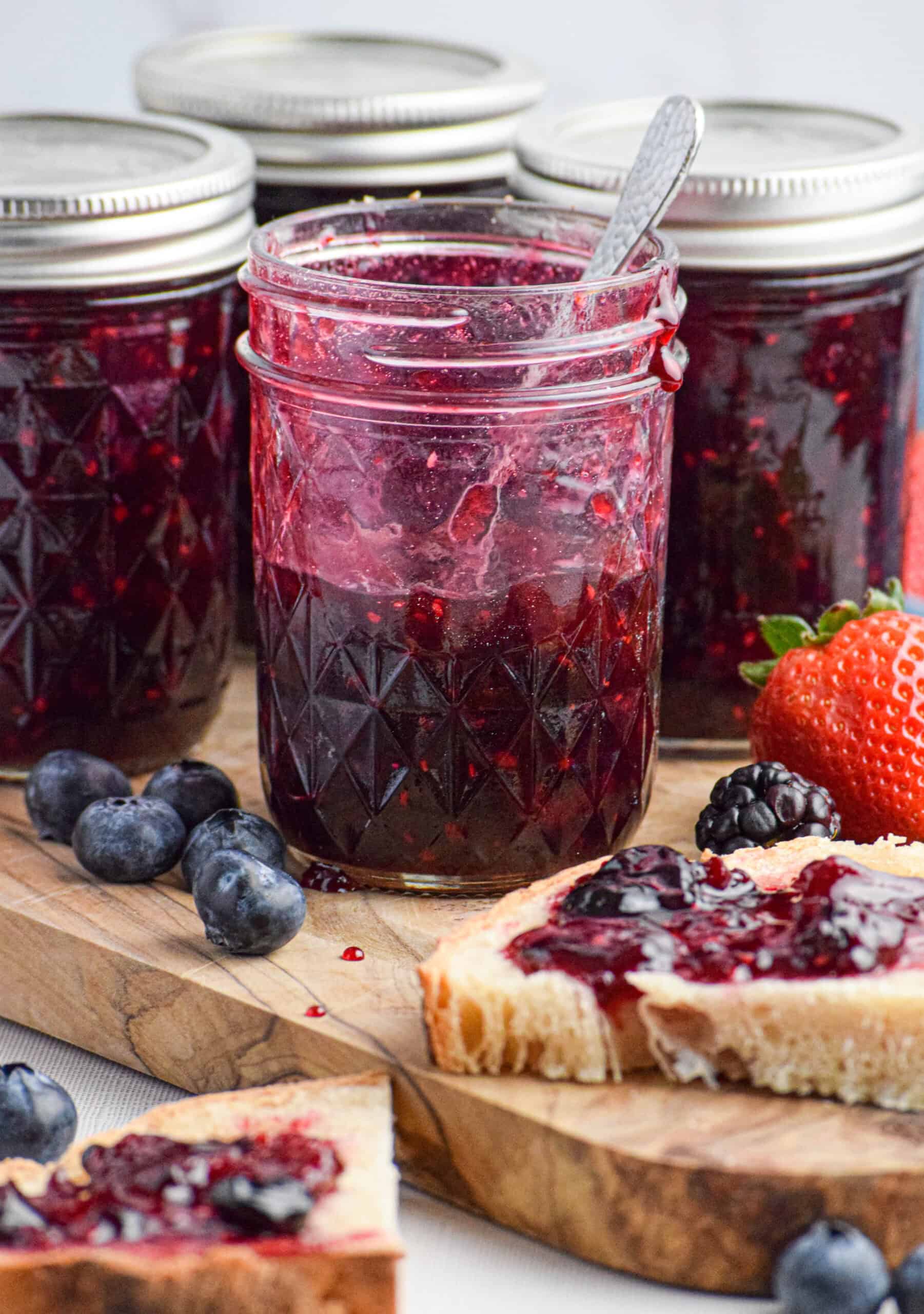 An open, half used jar of homemade berry preserves is sitting on a wood cutting board next to other sealed jars of jam, fresh berries and bread topped with jam. 