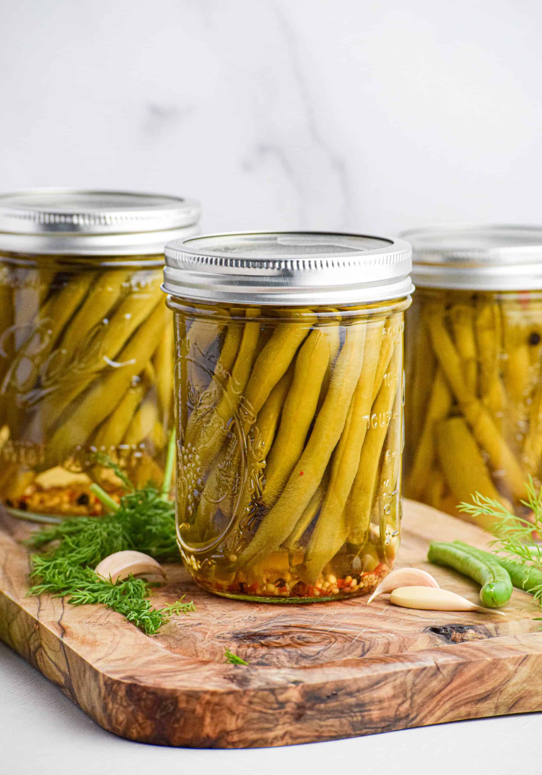 A mason jar of the best spicy dilly pickled green beans is sitting on a wood cutting board next to other jars of pickled, fresh dill and garlic cloves. 