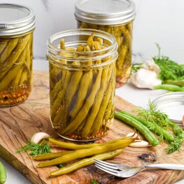 An open jar of pickled green beans sits on a wooden cutting board.