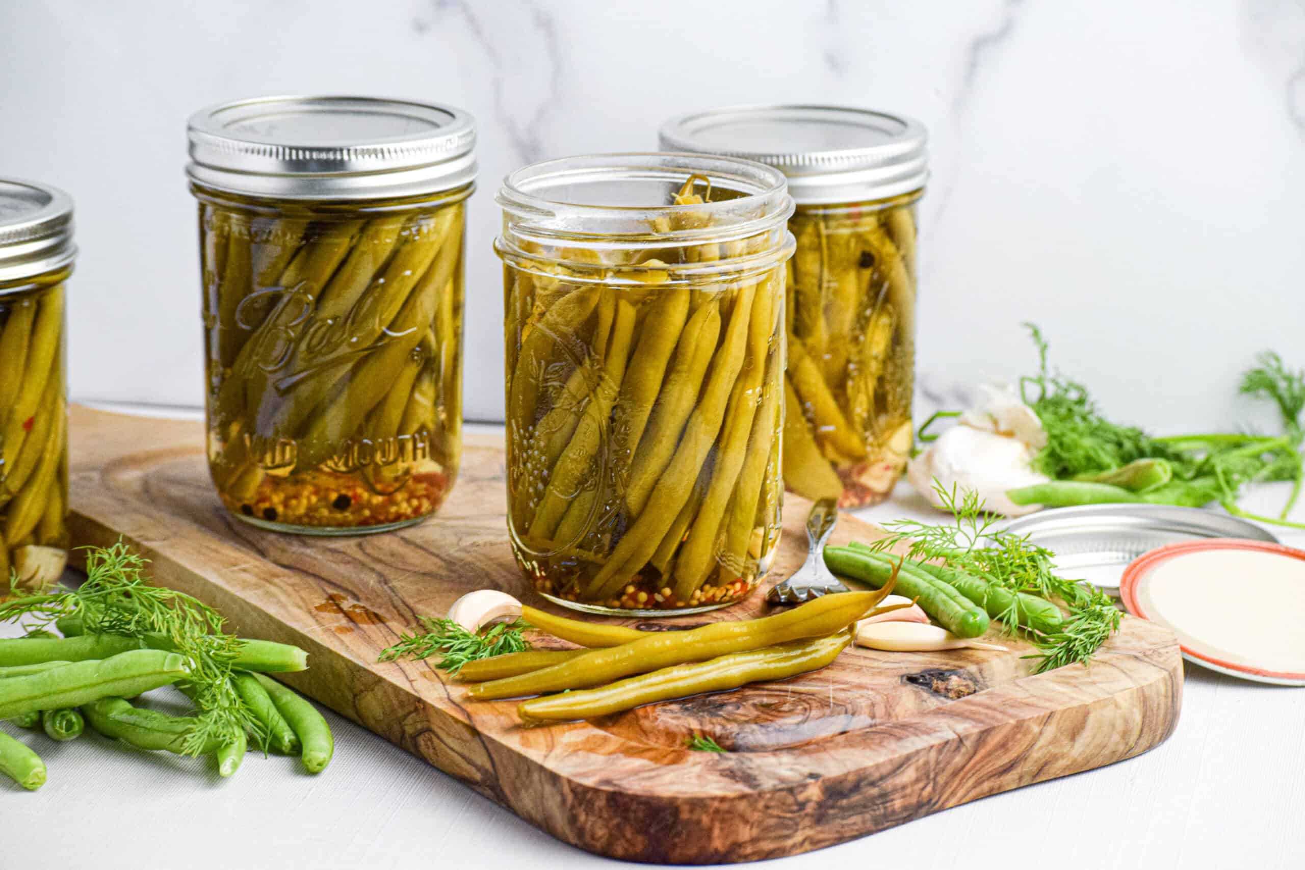 Several jars of the best homemade pickled green beans are sitting on a wooden cutting board, one is open and there are several pickled beans out along with sprigs of fresh dill and garlic cloves. 