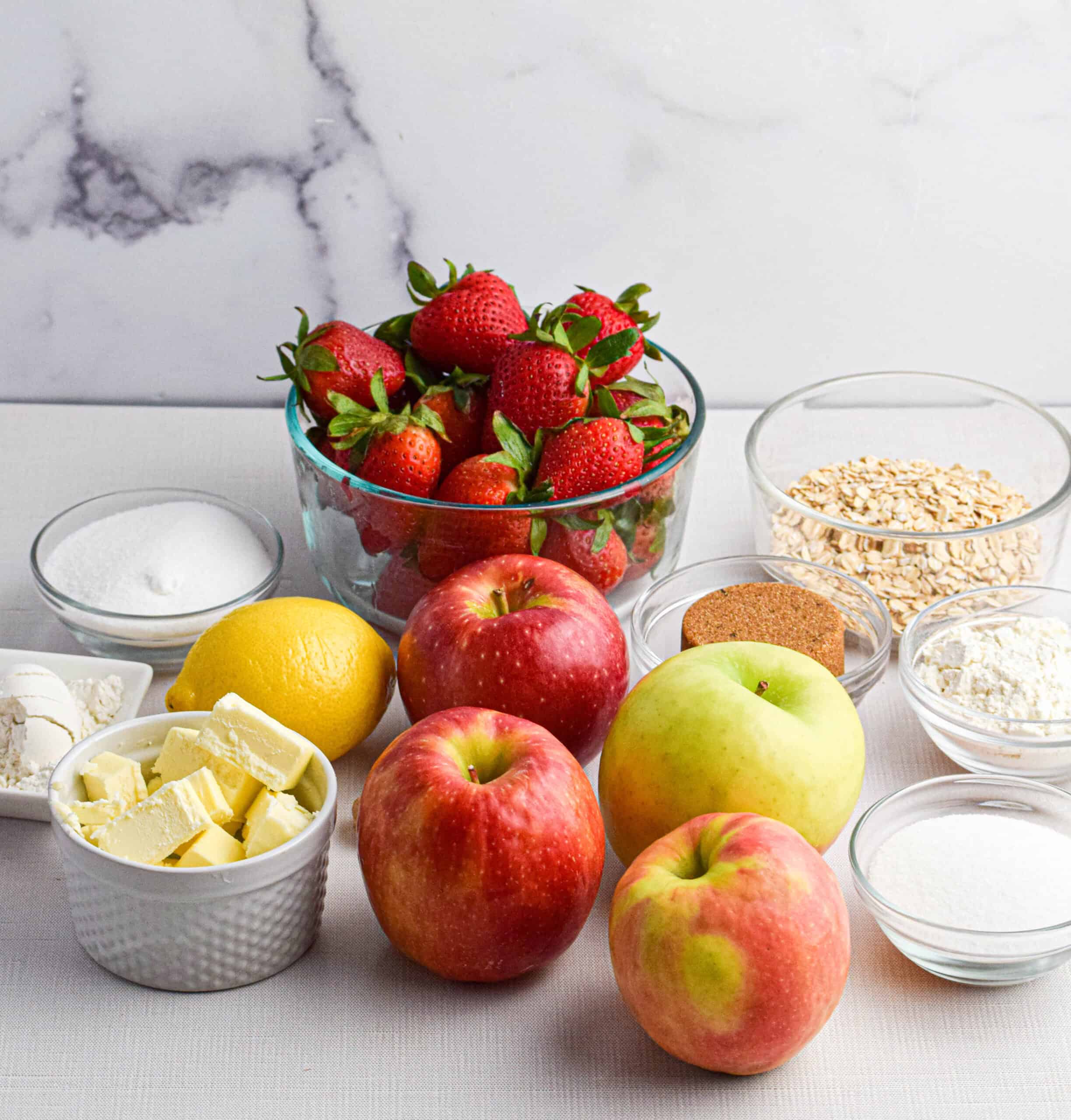 Ingredients for a homemade crumble with apples and strawberries is laid out on a white surface, fresh apples and strawberries, oats, sugar, flour, butter, and a lemon. 