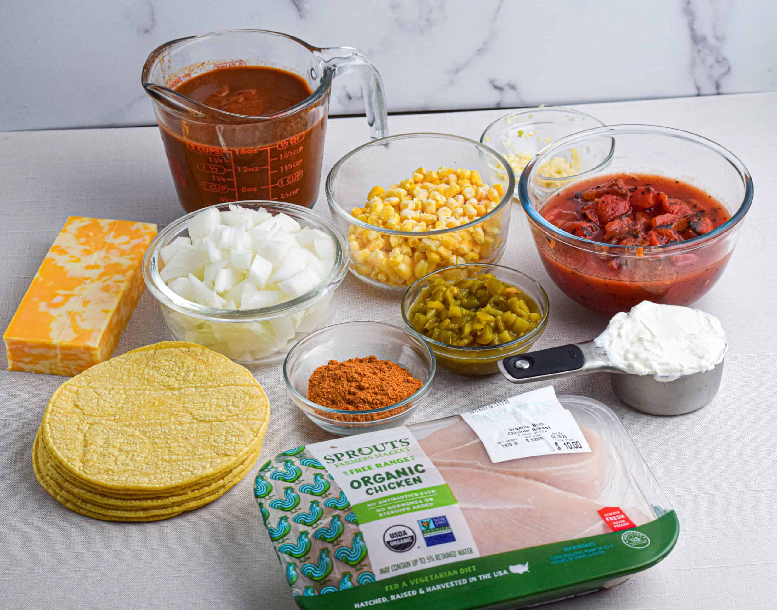 Ingredients for making easy crock pot chicken enchilada casserole are laid out including: chicken breasts, corn tortillas, sour cream, green chiles, taco seasoning, diced tomatoes, corn, diced onion, garlic, enchilada sauce and a block of cheese. 