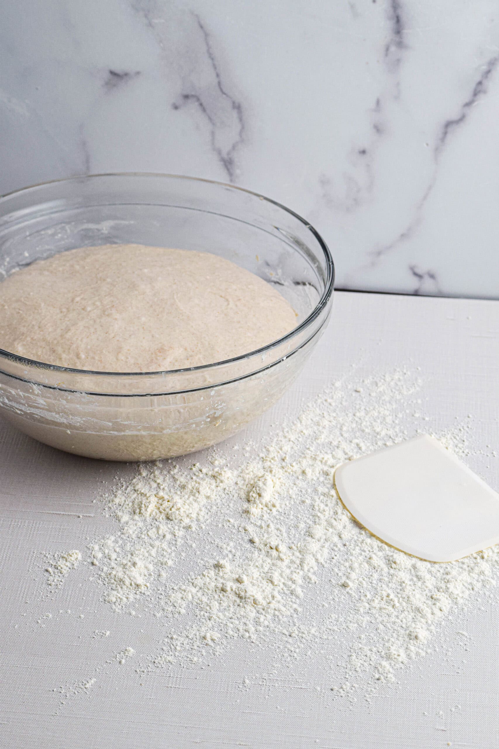 A glass bowl with risen sourdough dough by a floured surface and white dough scraper. 