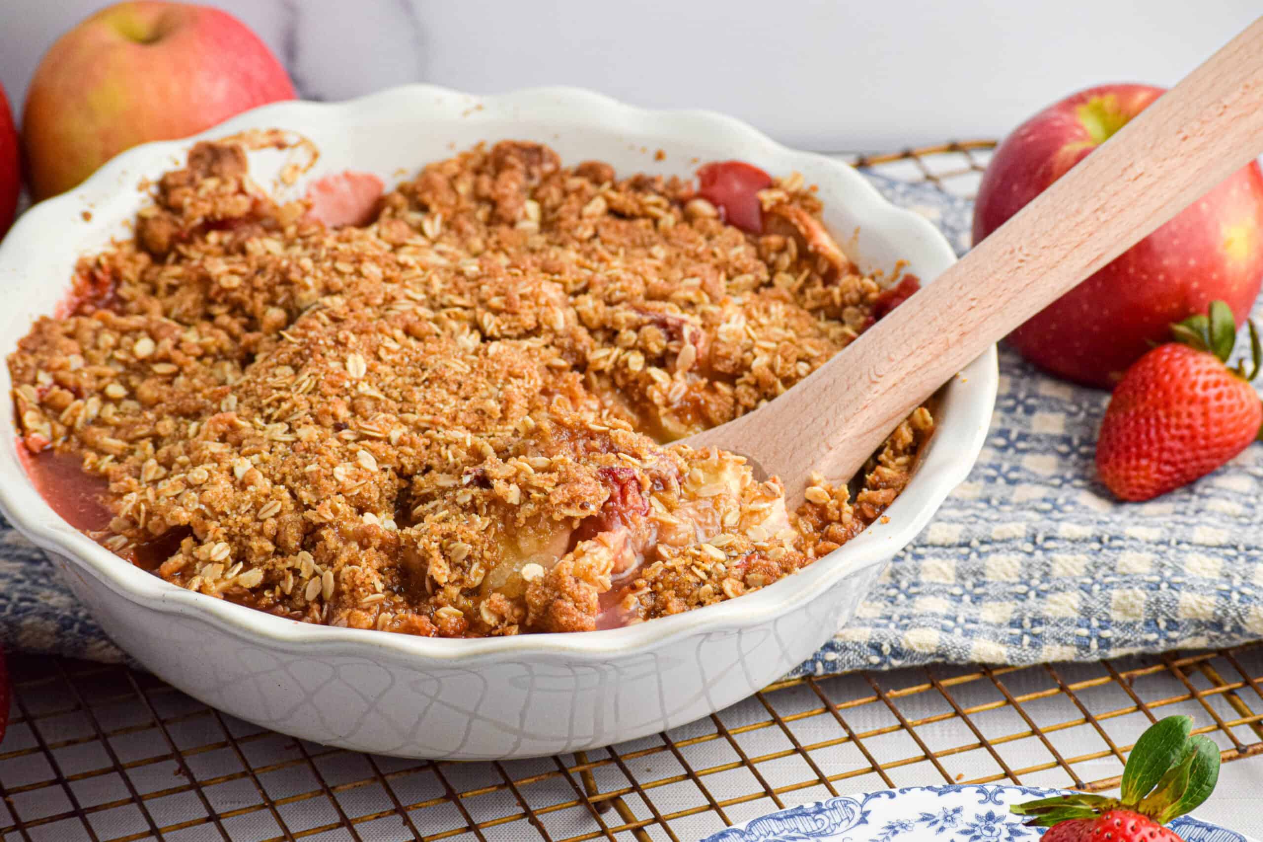 A white round pie dish is full of baked strawberry apple crumble with a wooden serving spoon. 