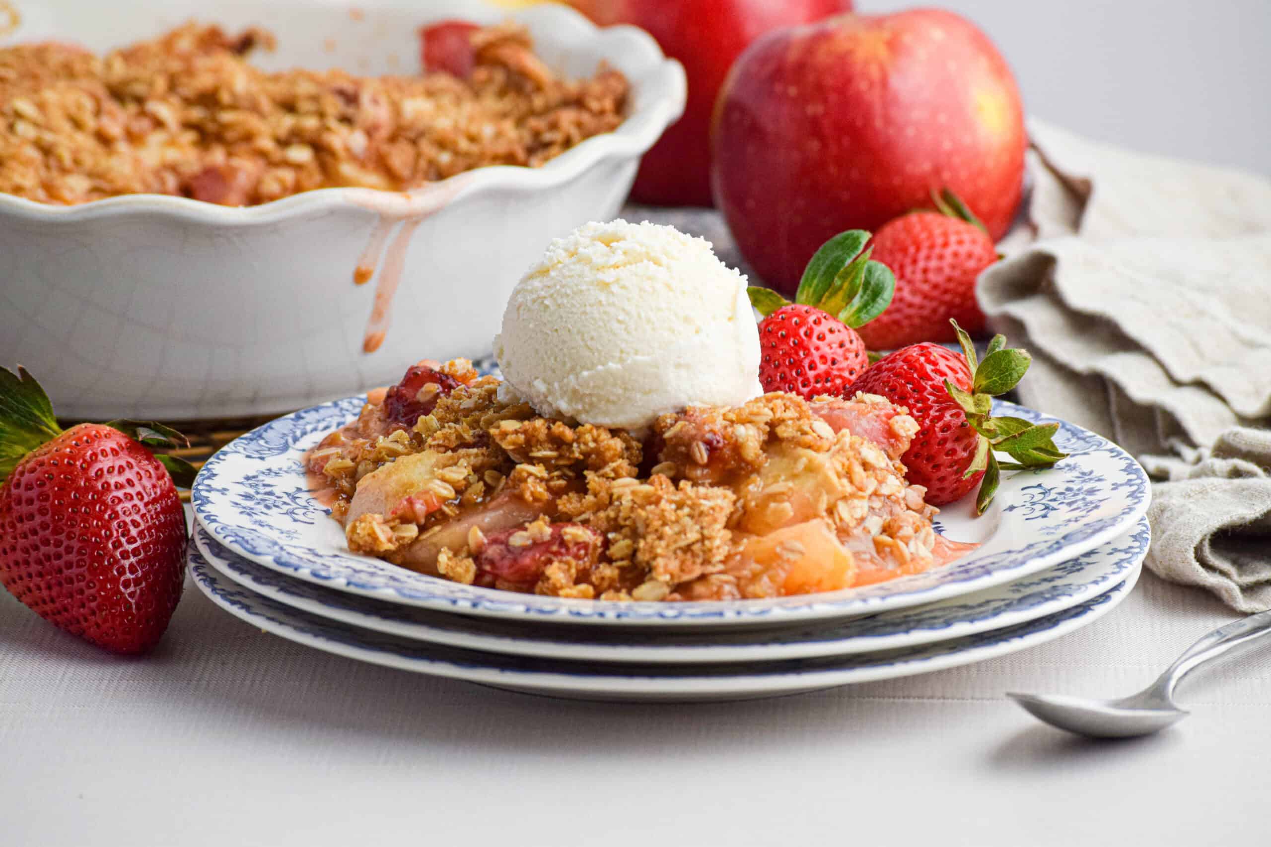 Serving of strawberry apple crumble with a scoop of vanilla ice cream surrounded by strawberries and apples. 