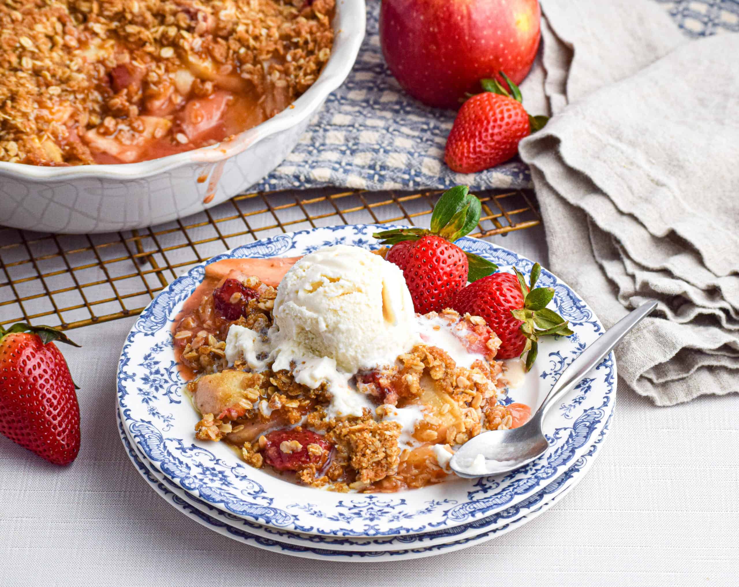 A serving of strawberry and apple crisp with a crumble topping and a scoop of vanilla ice cream is on a stack of blue and white plates surrounding by fresh strawberries, and apple and a serving dish of crumble. 