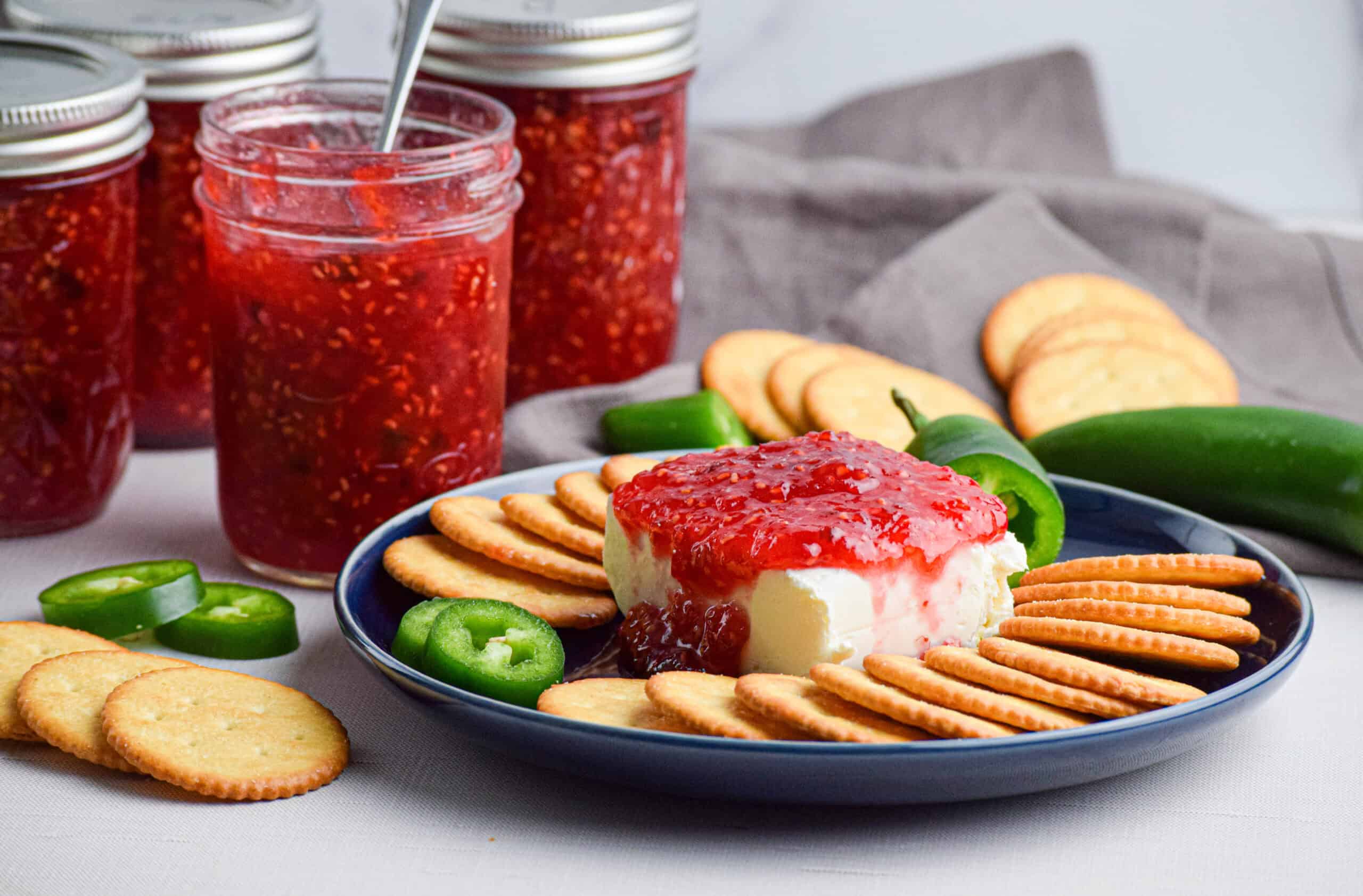 Jars of raspberry jalapeno pepper jelly and a plate with cream cheese topped with raspberry jam with jalapenos and crackers.
