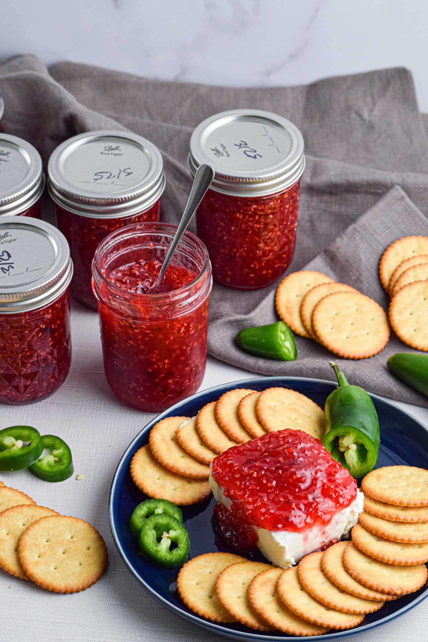 Several jars of raspberry jalapeno jam and a plate with a block of cream cheese topped with the preserves and surrounded by crackers and sliced fresh jalapenos.