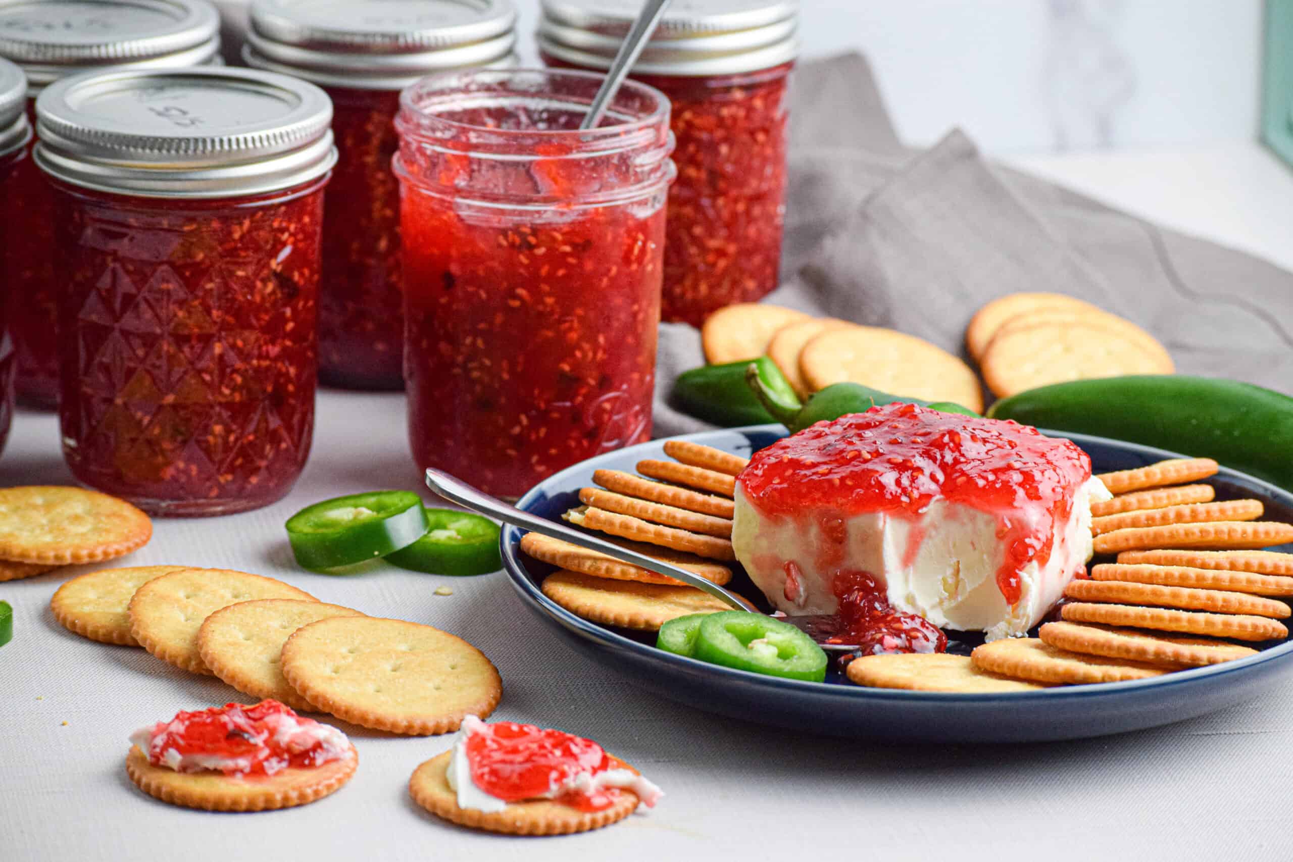 A plate with the easy appetizer of cream cheese topped with homemade jalapeno raspberry preserves served on crackers with several jars of jam that have been water bath canned.