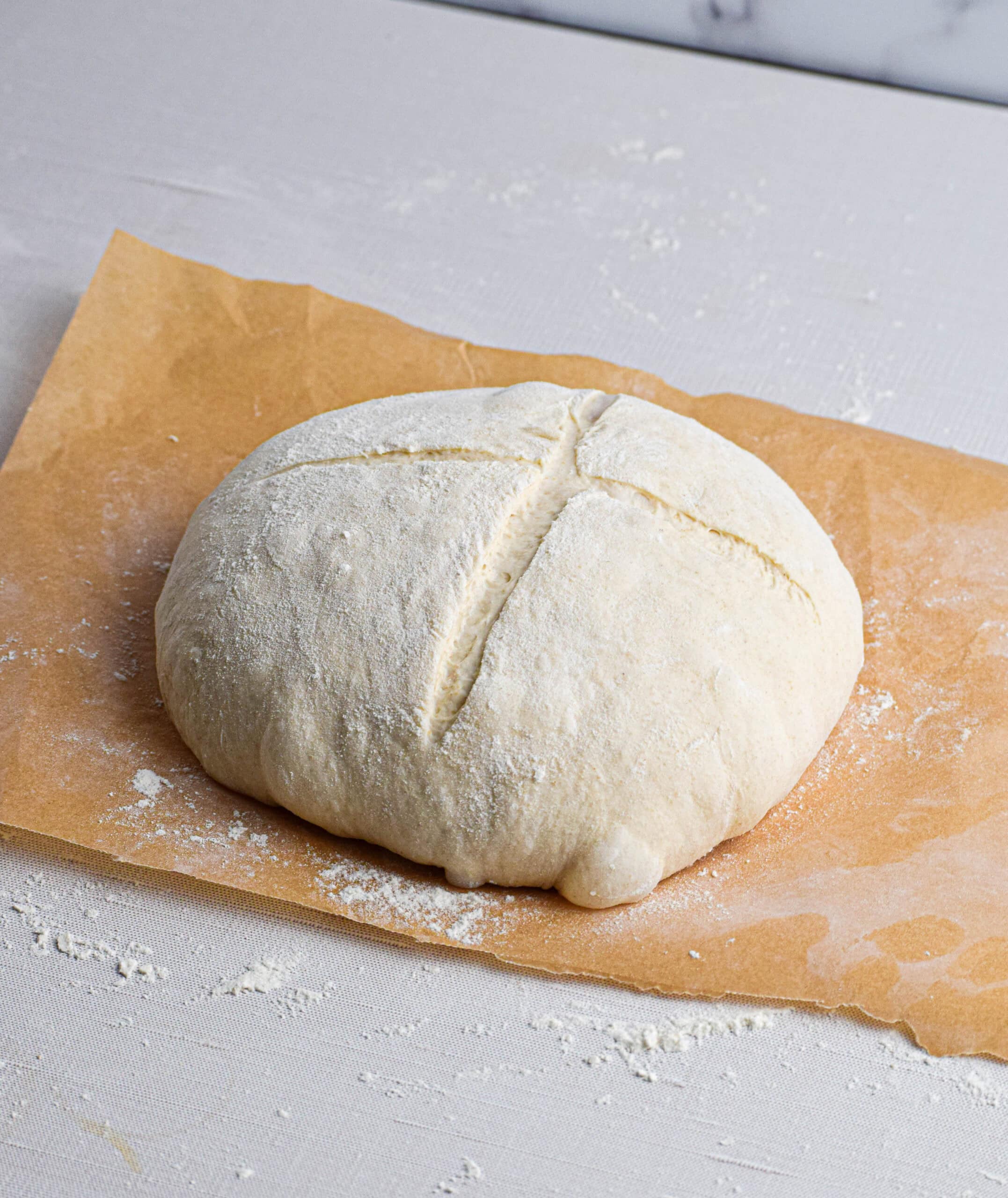 The shaped and risen dough has been placed on a floured sheet of parchment paper with a cross shape scored onto the top. 