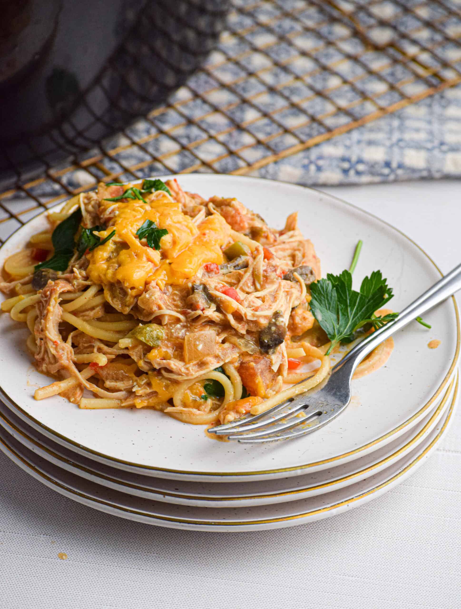 A close-up of a plate of the best creamy slow cooker chicken spaghetti, topped with melted cheddar cheese and garnished with fresh parsley; the plate rests on a stack of dishes with a silver fork placed beside the pasta and a cooling rack and patterned towel are blurred in the background.