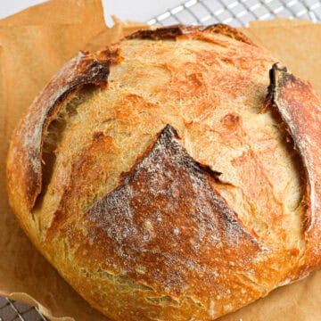 A beautiful sourdough boule baked to golden brown, crispy crust perfection sits on a piece of parchment paper on a cooling rack.