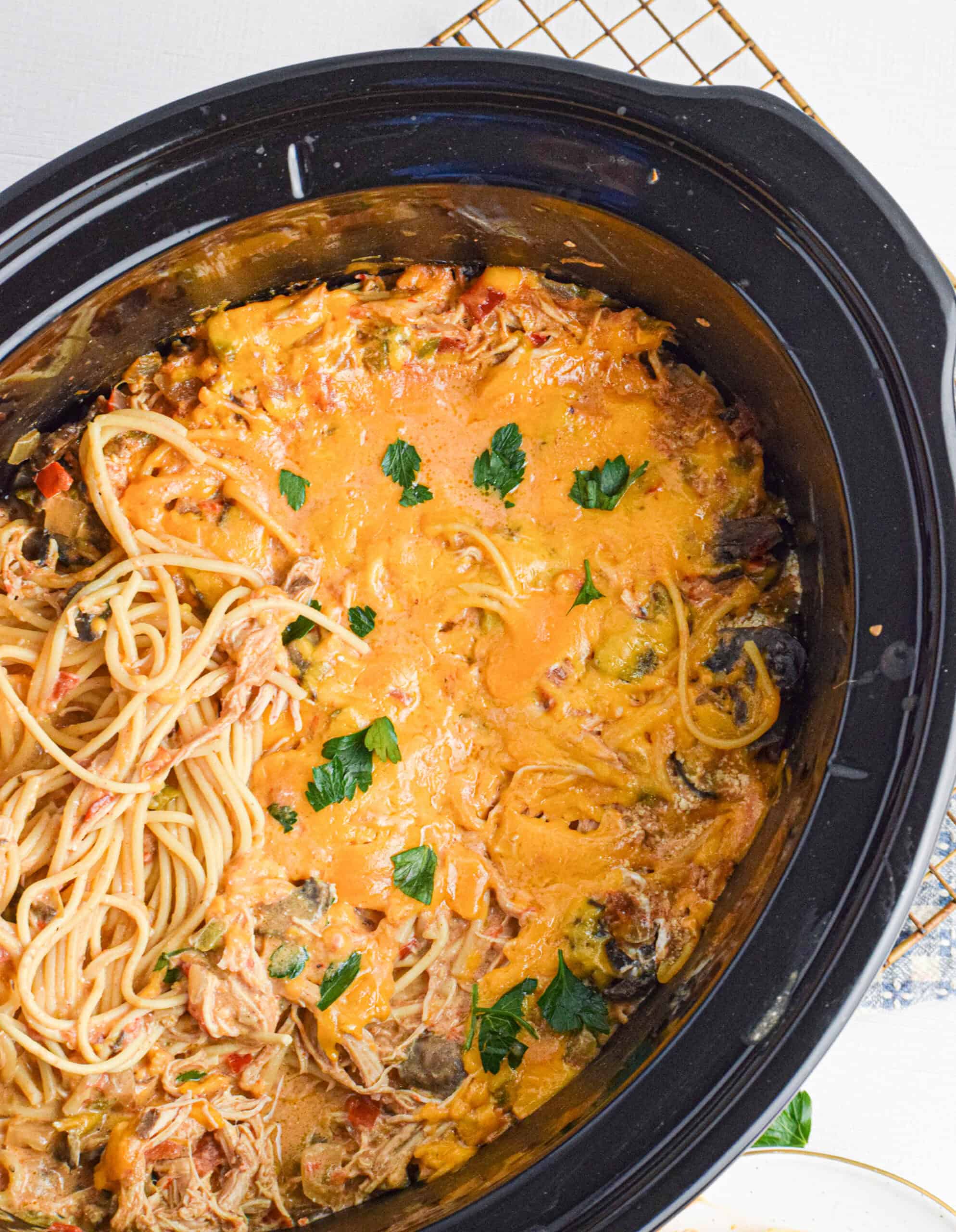 Overhead view of a slow cooker filled with cheesy crock pot chicken spaghetti. The dish is creamy and colorful with shredded chicken, spaghetti noodles, melted cheddar cheese, and garnished with fresh parsley. One side of the slow cooker shows the pasta being scooped out.