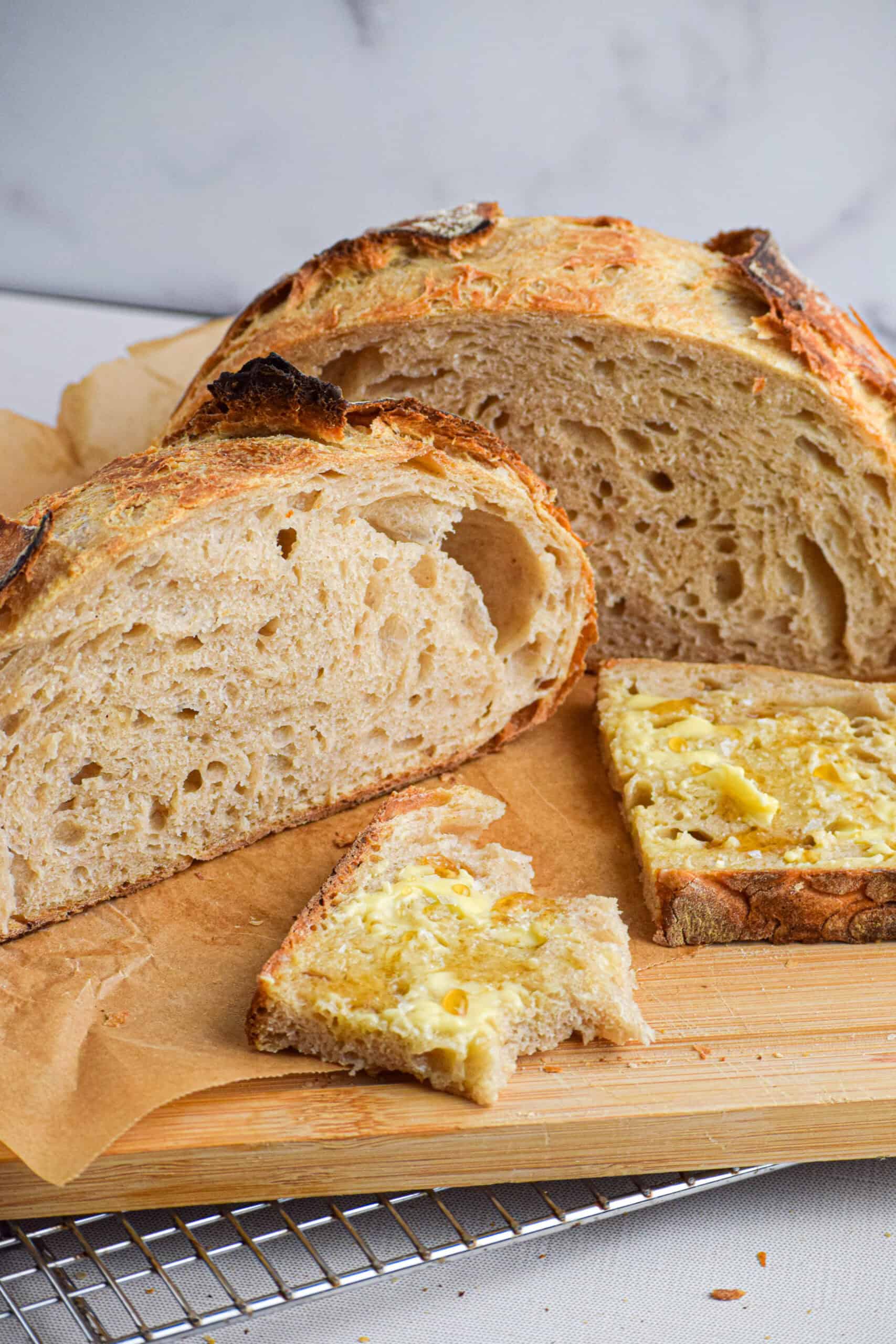 A loaf of homemade sourdough bread cut in half and sitting on a wood cutting board with a slice covered but butter and honey. 