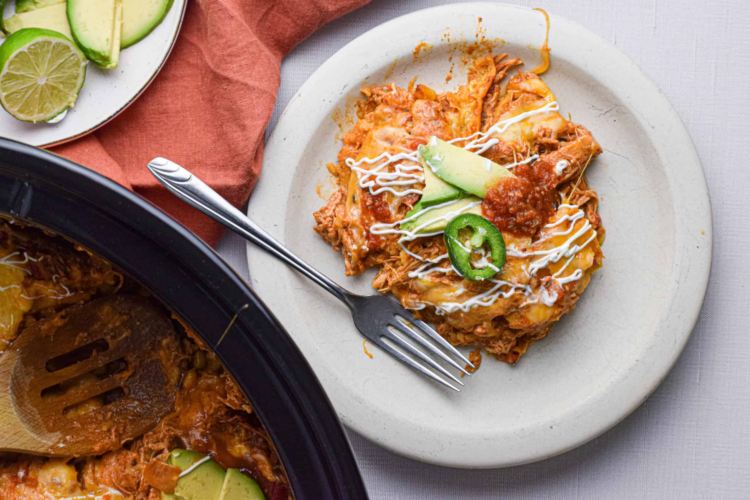 A serving of chicken enchilada casserole topped with a jalapeno and avocado slice, sour cream and salsa is on a plate with a fork next to a large slow cooker bowl. 