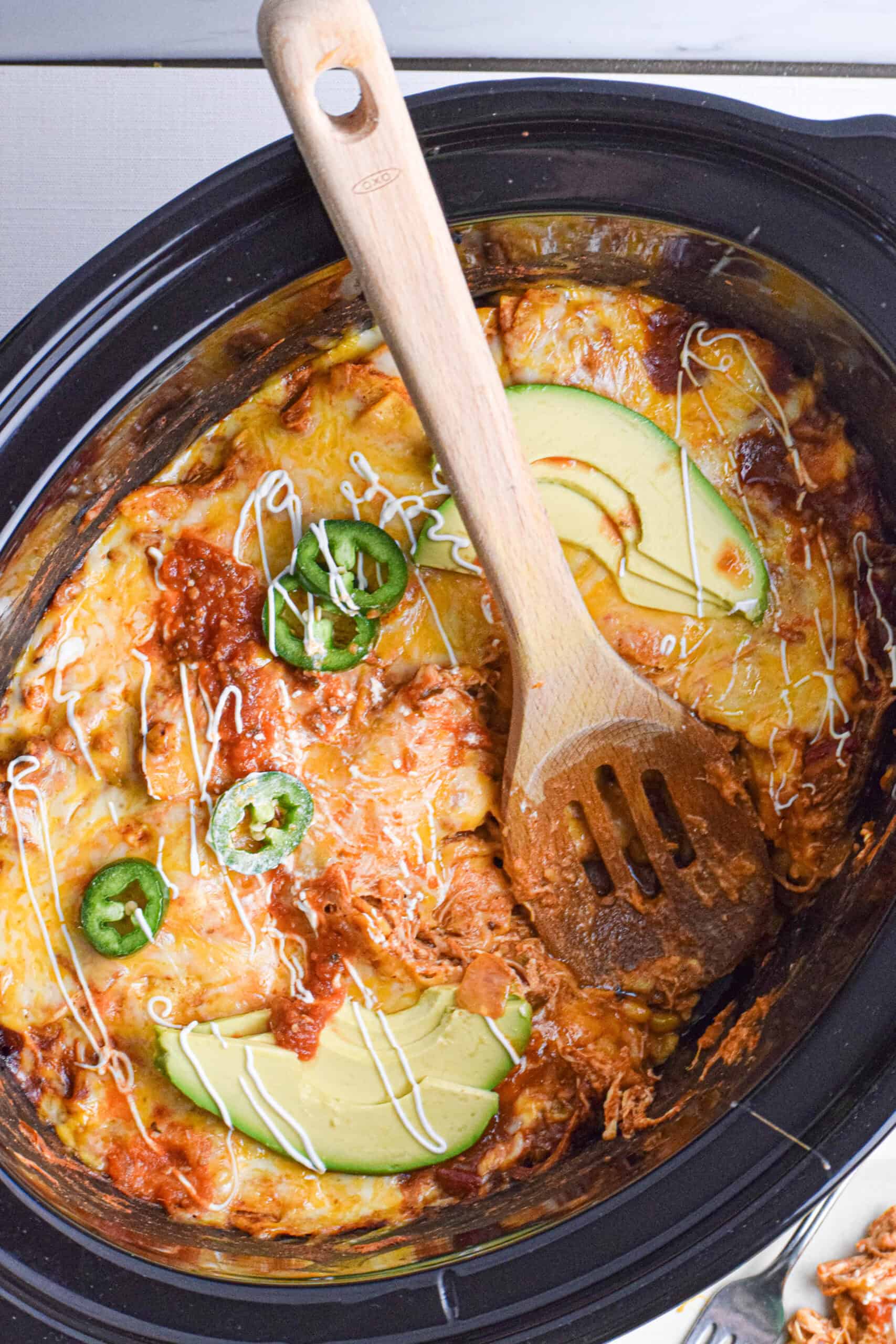 Overhead shot of a enchilada casserole in the bowl of a slow cooker topped with melted shredded cheese, sliced peppers, avocados, sour cream and salsa with a wooden spoon taking a serving. 