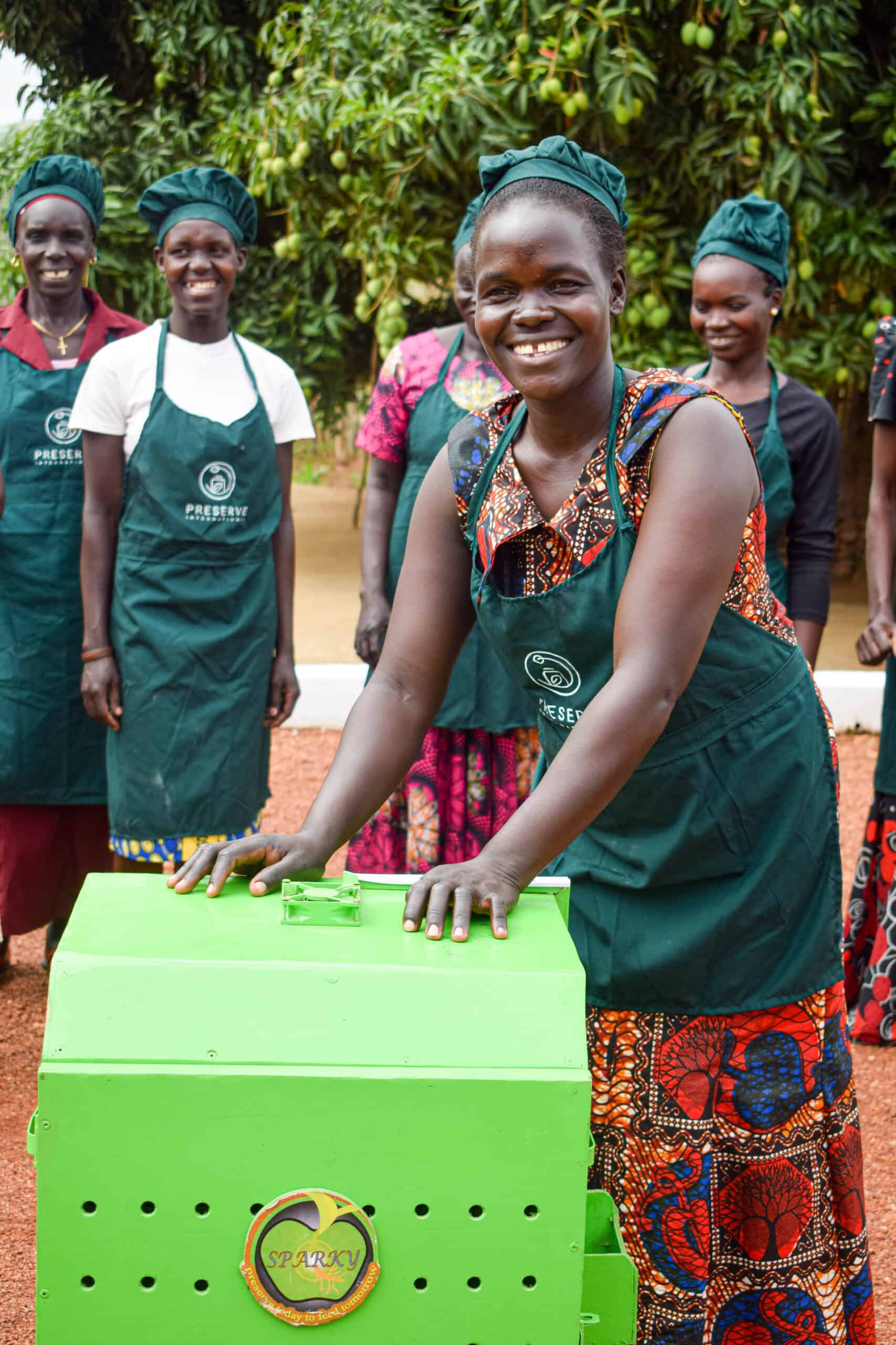 Smiling women wearing Preserve International aprons stand together outdoors. One woman stands at the front with her hands on a bright green Sparky Dryer, a solar-powered food dehydrator used for preserving crops.