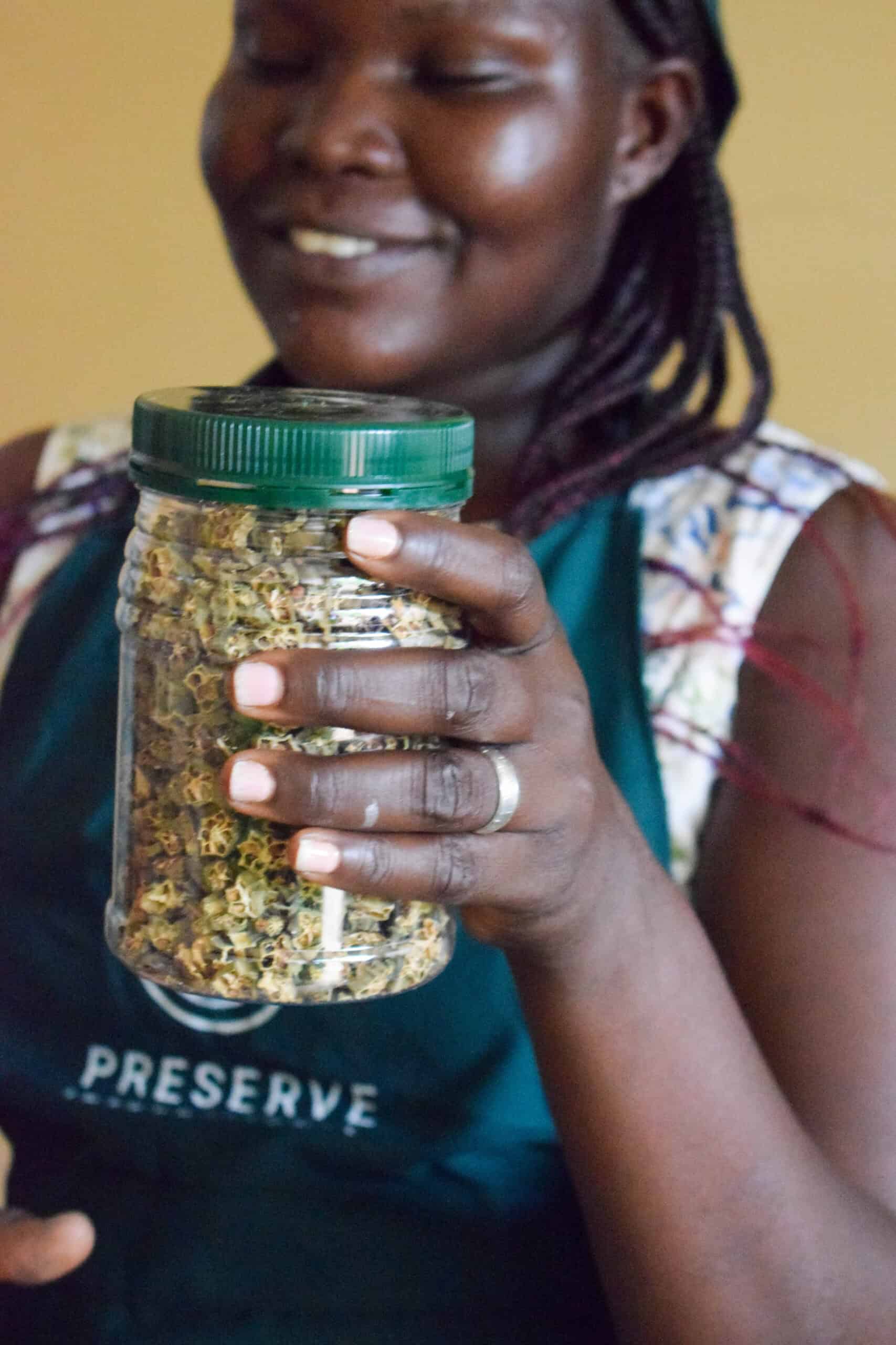A woman wearing a Preserve International apron holds up a clear jar filled with safely dried okra, showcasing preserved produce from training in food dehydration techniques.
