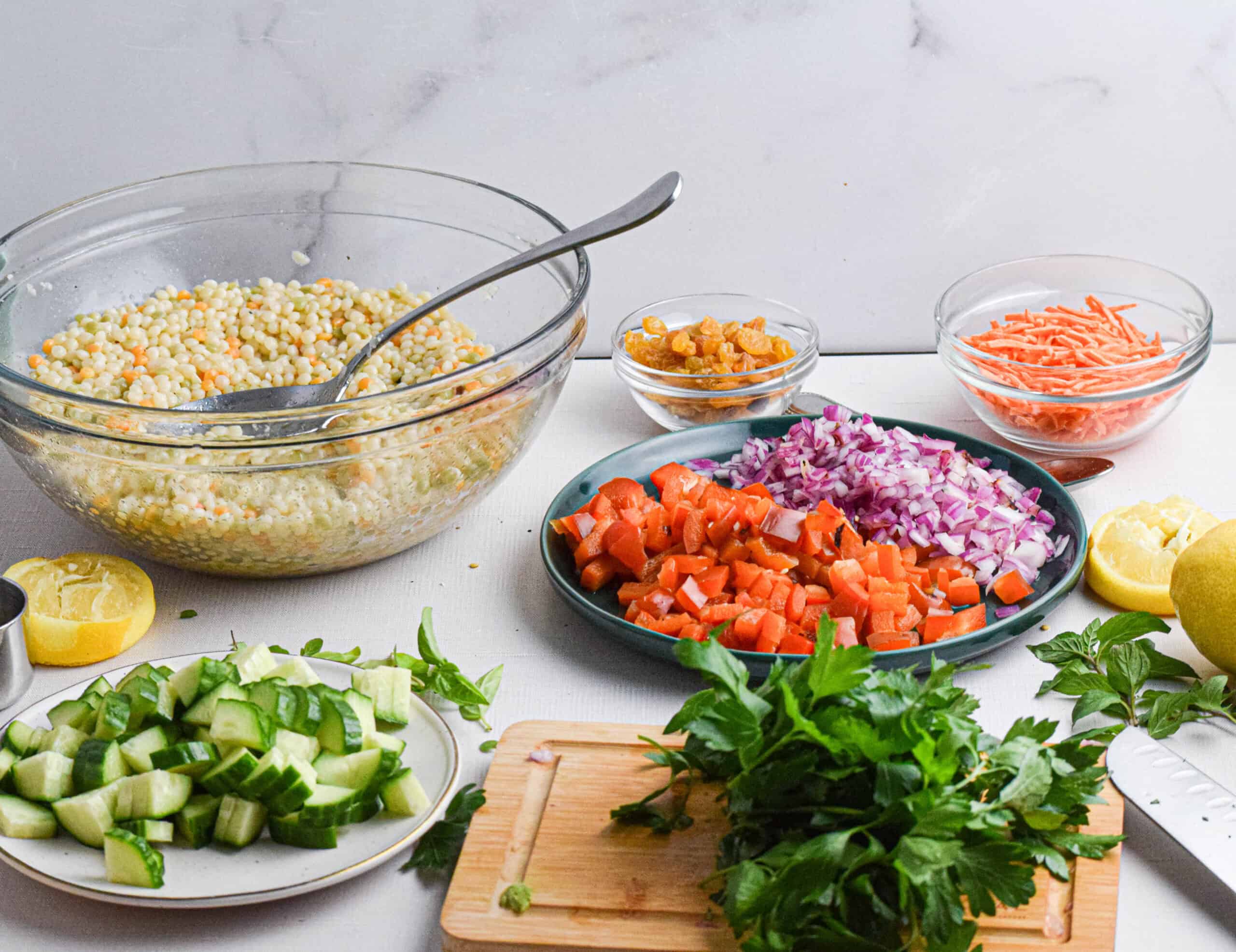 Chopped vegetables, herbs, and lemon on a counter with a large glass bowl of couscous salad being prepared for a fresh and flavorful meal.