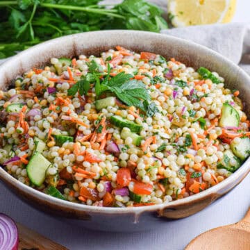 A bowl of couscous salad with chopped vegetables, garnished with parsley, sits on a table next to a wooden spoon, fresh parsley, lemon wedges, and sliced red onion.
