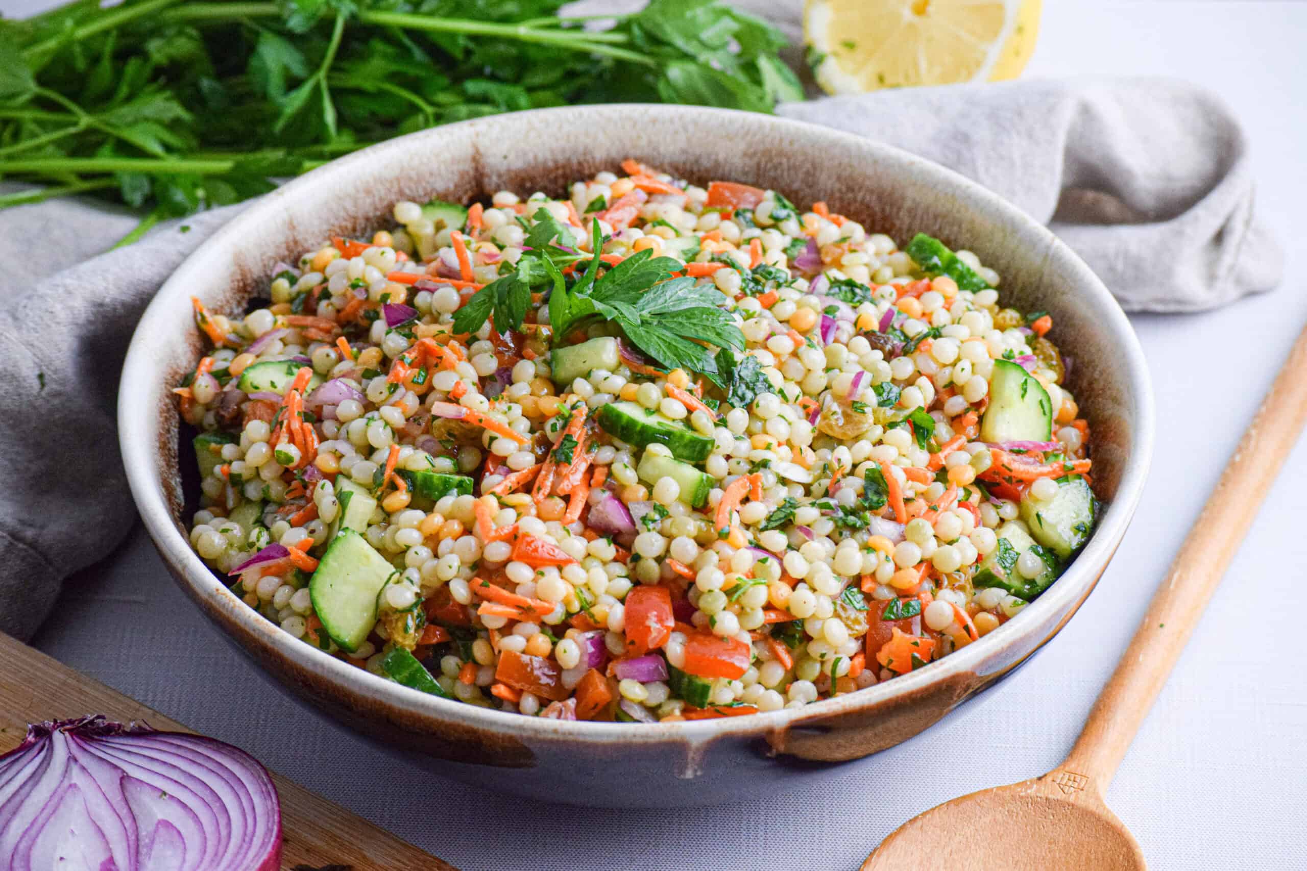 A bowl of couscous salad with chopped vegetables, garnished with parsley, sits on a table next to a wooden spoon, fresh parsley, lemon wedges, and sliced red onion.