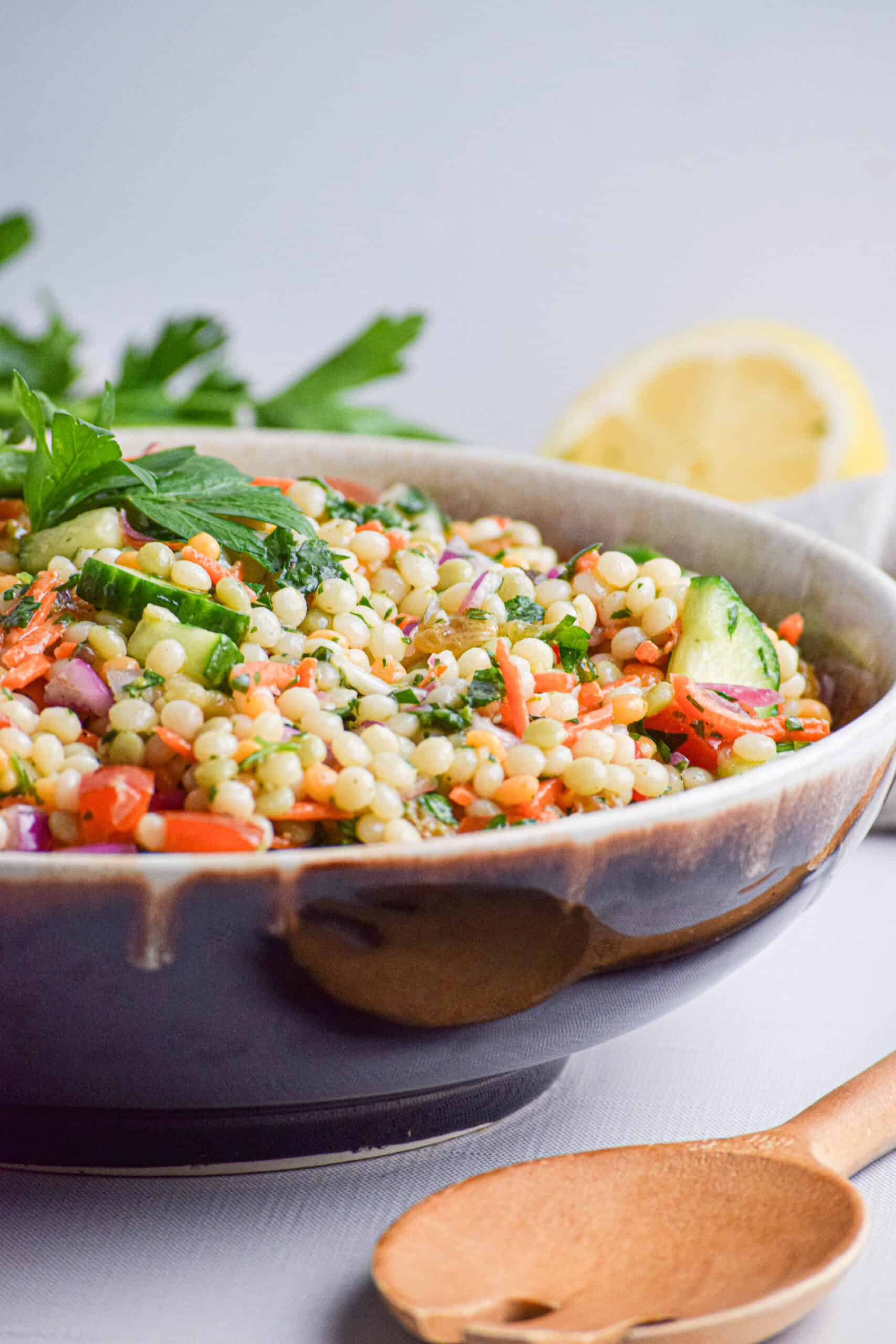 A vibrant couscous salad with chopped vegetables and herbs, garnished with parsley, sits in a bowl, accompanied by a wooden spoon and a lemon wedge in the background.