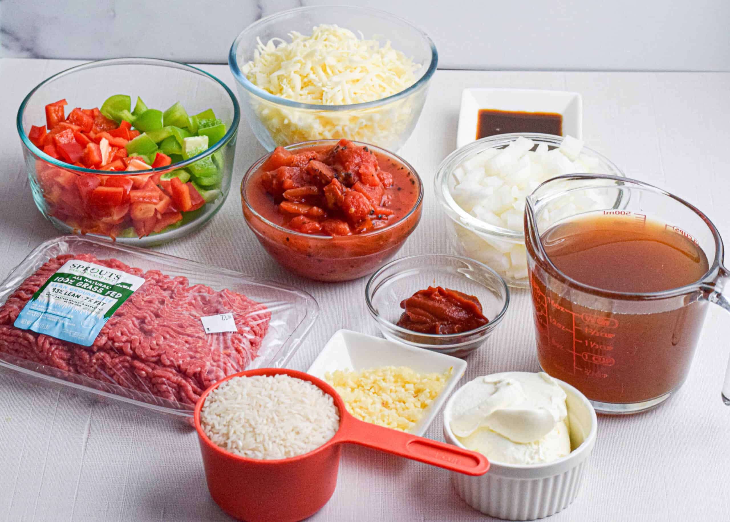 A variety of ingredients for stuffed pepper casserole on a white surface, including ground beef, rice, cheese, chopped vegetables, broth, and seasonings in bowls and containers.