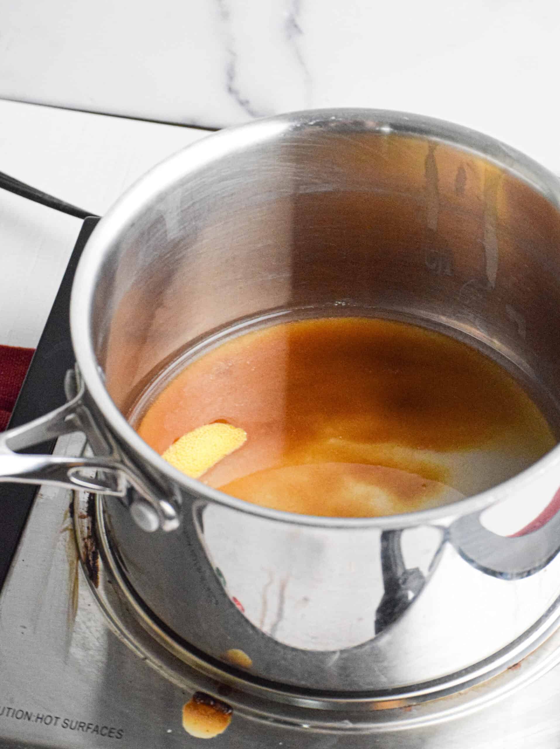 A stainless steel pot on a stove contains brown liquid and a floating lemon peel, possibly simmering blueberry compote. Some liquid has dripped onto the hot plate surface.
