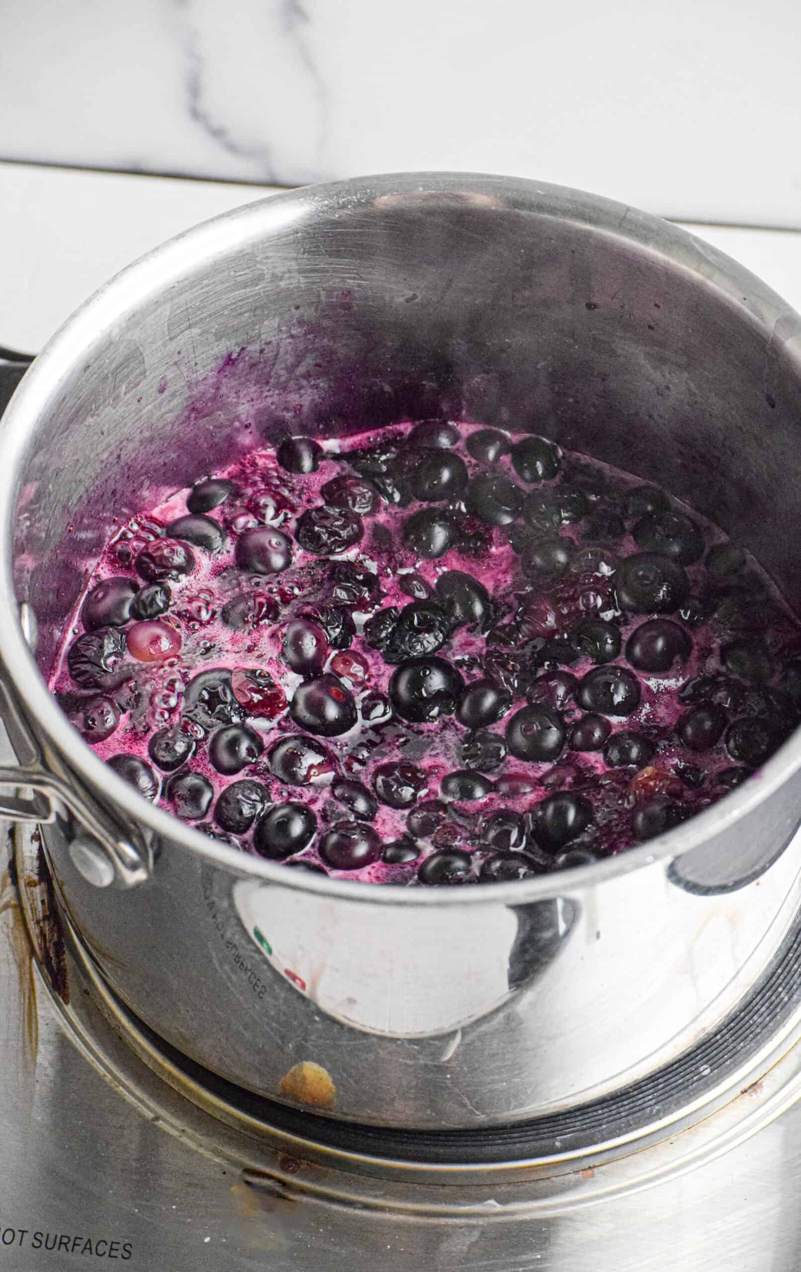 A stainless steel pot on a stove contains simmering blueberries in a purple liquid, creating a rich blueberry compote.