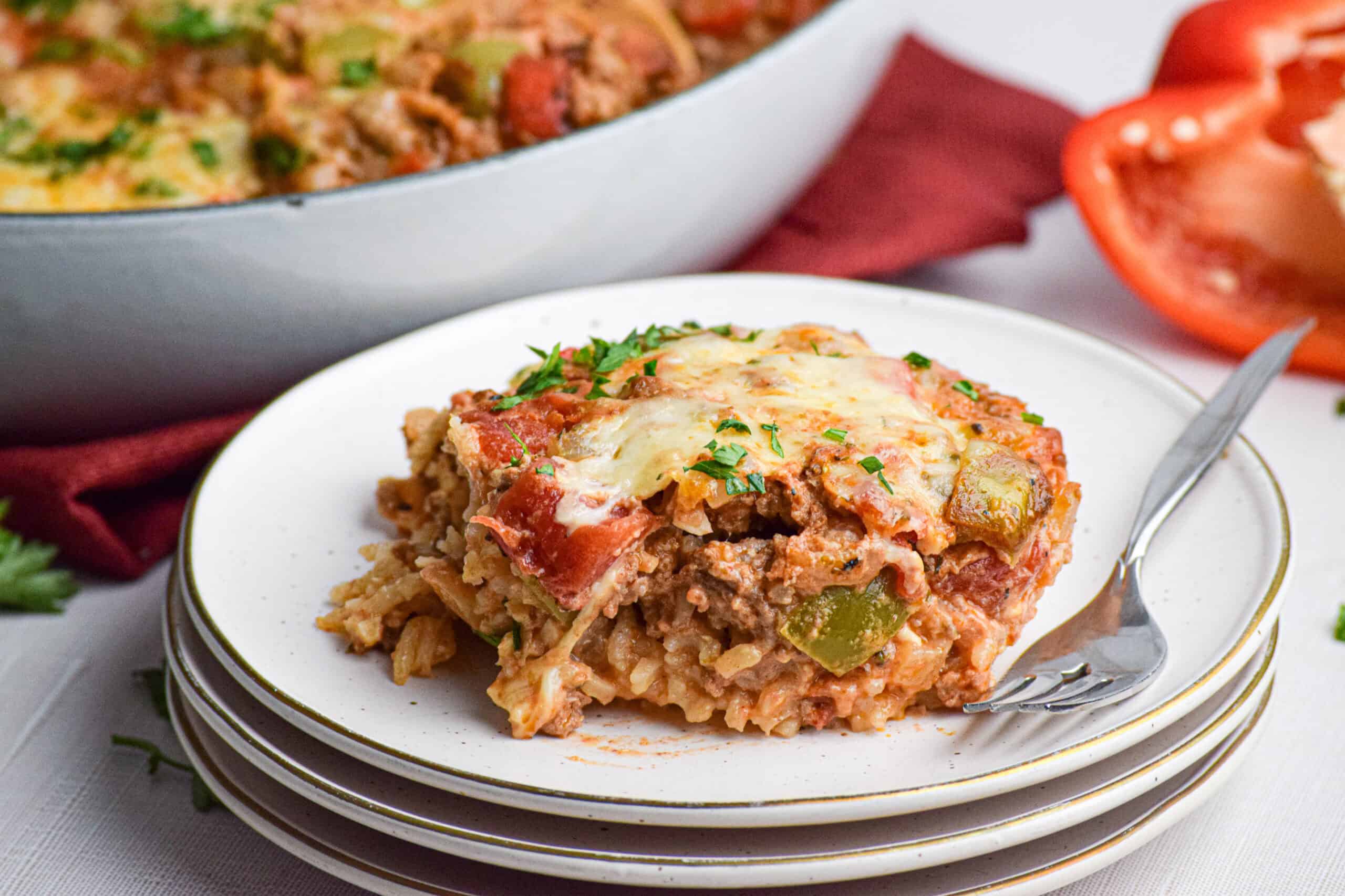 A serving of stuffed pepper casserole with rice, ground meat, bell peppers, and melted cheese on a white plate with a fork, next to a sliced bell pepper.