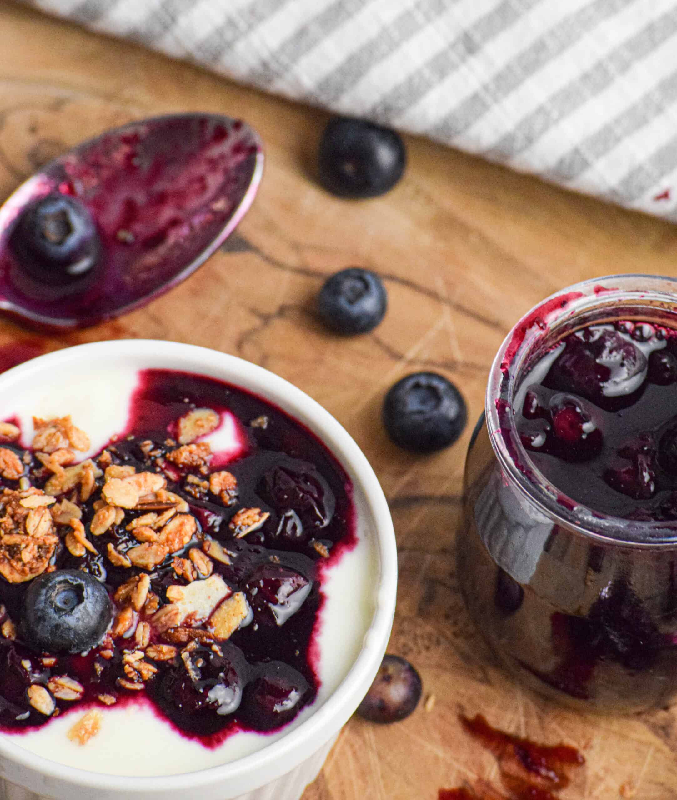 A bowl of yogurt topped with granola, blueberries, and blueberry compote sits next to a jar of blueberry sauce and scattered blueberries on a wooden surface.