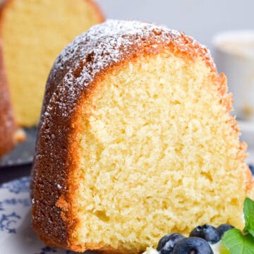 A slice of sour cream pound cake dusted with powdered sugar is served on a plate with whipped cream and fresh blueberries.