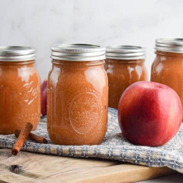 Four jars of homemade slow cooker applesauce with metal lids are displayed on a wooden surface alongside two whole apples, cinnamon sticks, and a blue cloth.