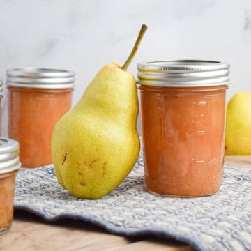 Four glass jars filled with homemade pear preserves sit on a blue textured cloth, with two whole pears placed nearby on a wooden surface.