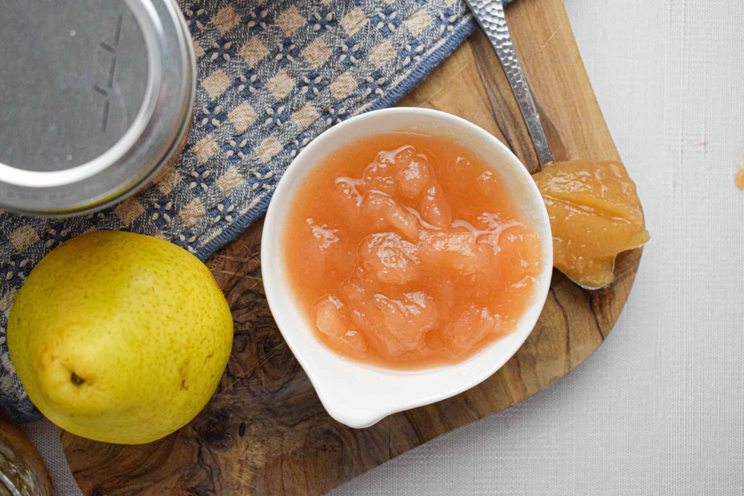 A bowl of pear preserves sits on a wooden board alongside a fresh pear, a spoon, a jar lid, and a blue checkered cloth.