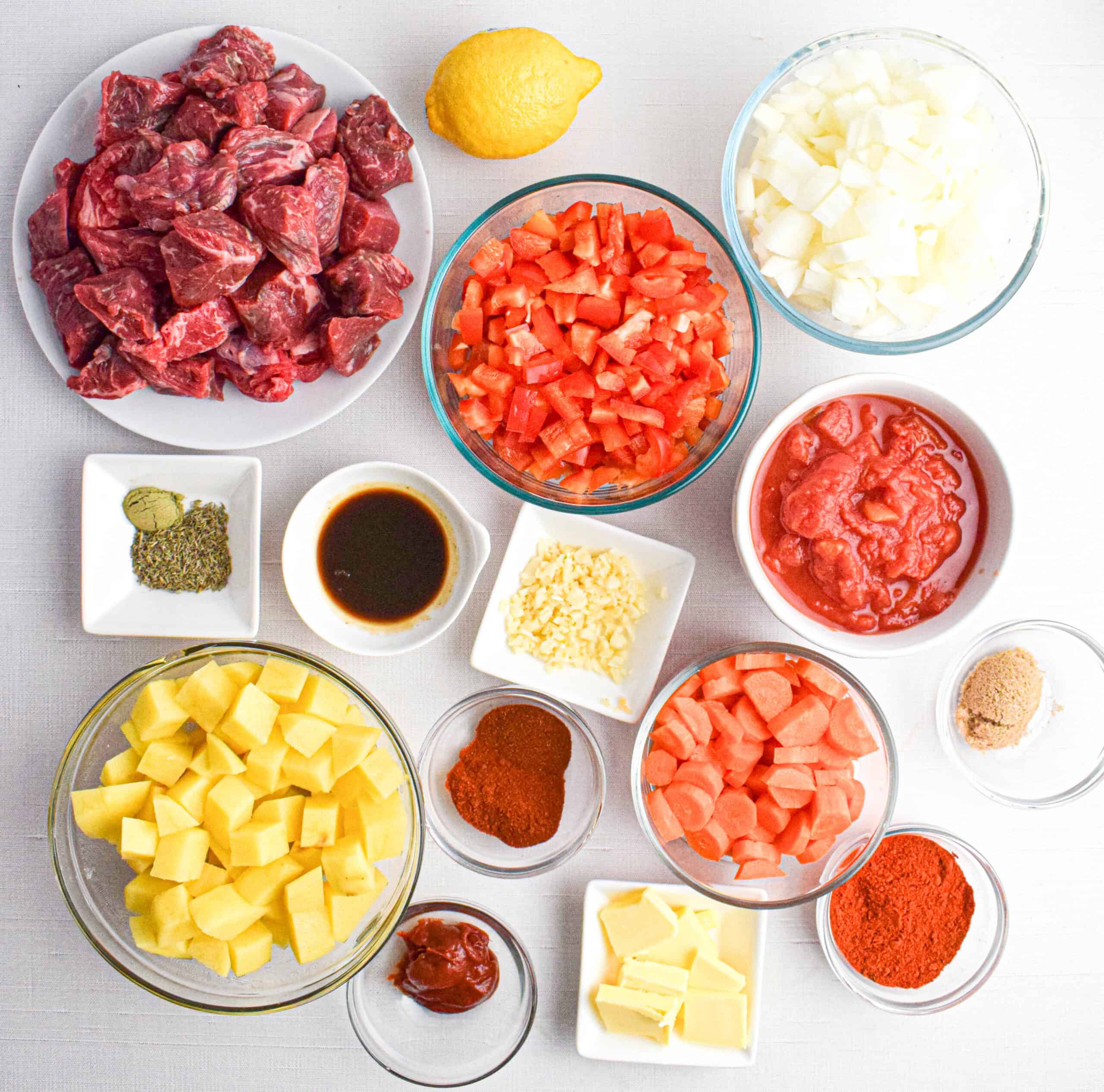 Assorted ingredients for Beef Goulash or stew, including diced beef, onions, tomatoes, potatoes, carrots, tomato paste, spices, garlic, brown sugar, lemon, and seasonings arranged in bowls on a white surface.