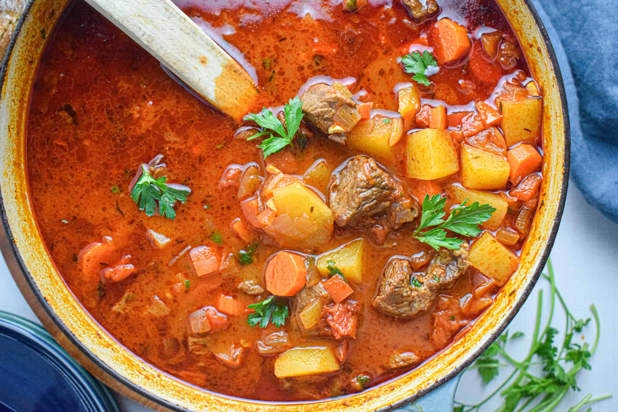 A pot of beef goulash stew with chunks of meat, carrots, potatoes, and garnished with parsley, shown with a wooden spoon.