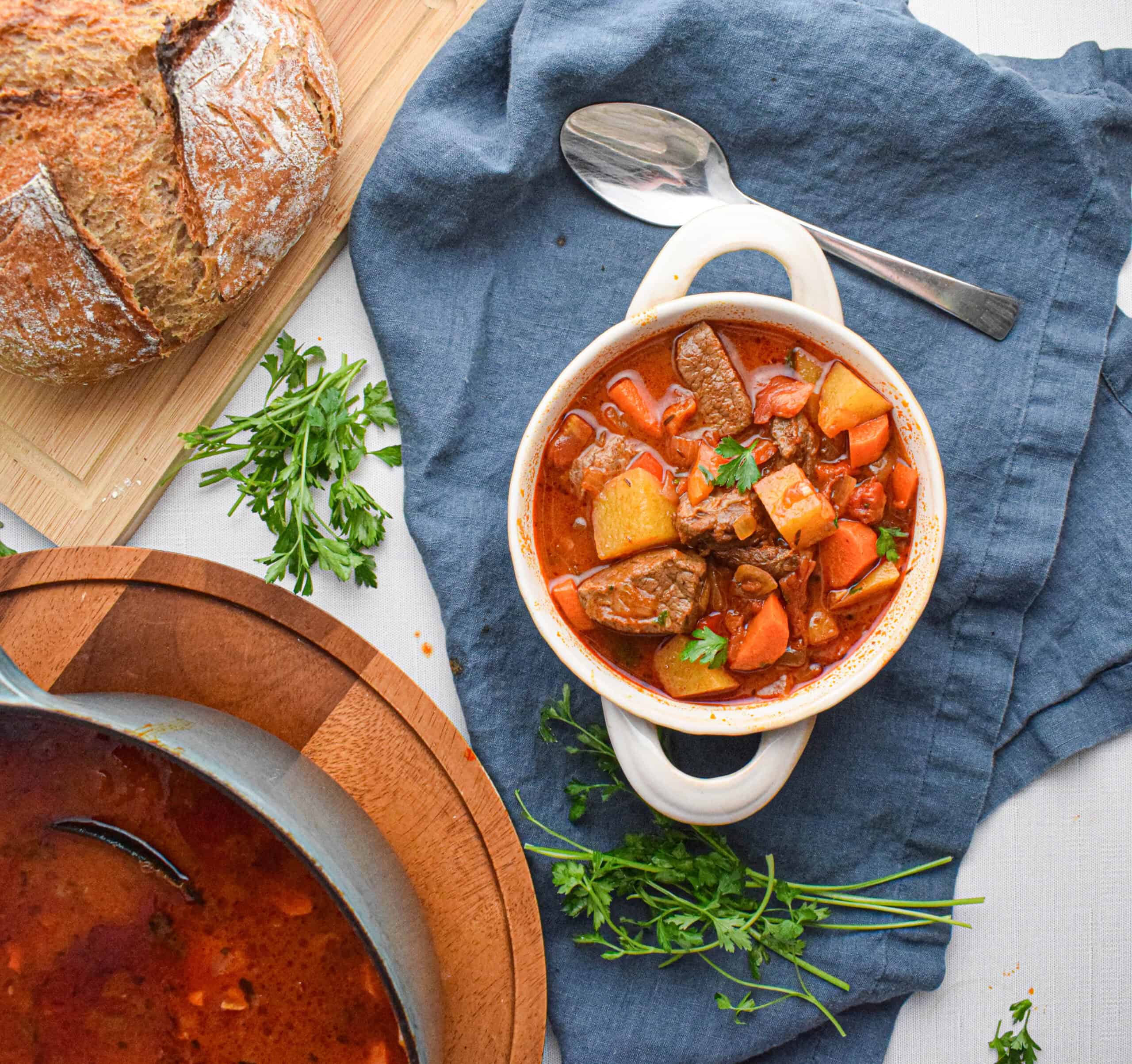 A bowl of hearty beef goulash with vegetables sits on a blue napkin next to a loaf of bread, fresh parsley, and a pot of stew on a light-colored table.