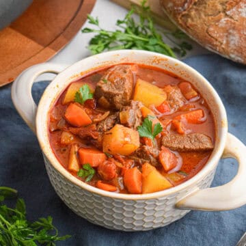 A bowl of beef goulash with carrots, potatoes, and parsley garnish sits on a blue cloth, accompanied by a loaf of bread and fresh parsley nearby.