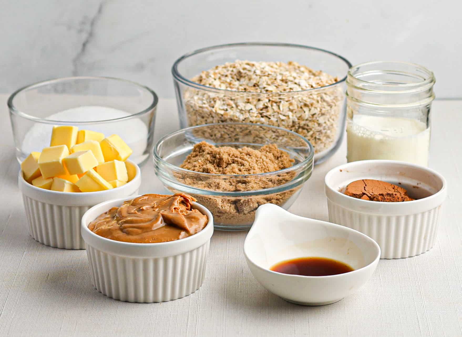 Several glass and ceramic bowls containing oats, cubed butter, sugar, brown sugar, peanut butter, cocoa powder, vanilla extract, and a small jar of milk are arranged on a white surface.