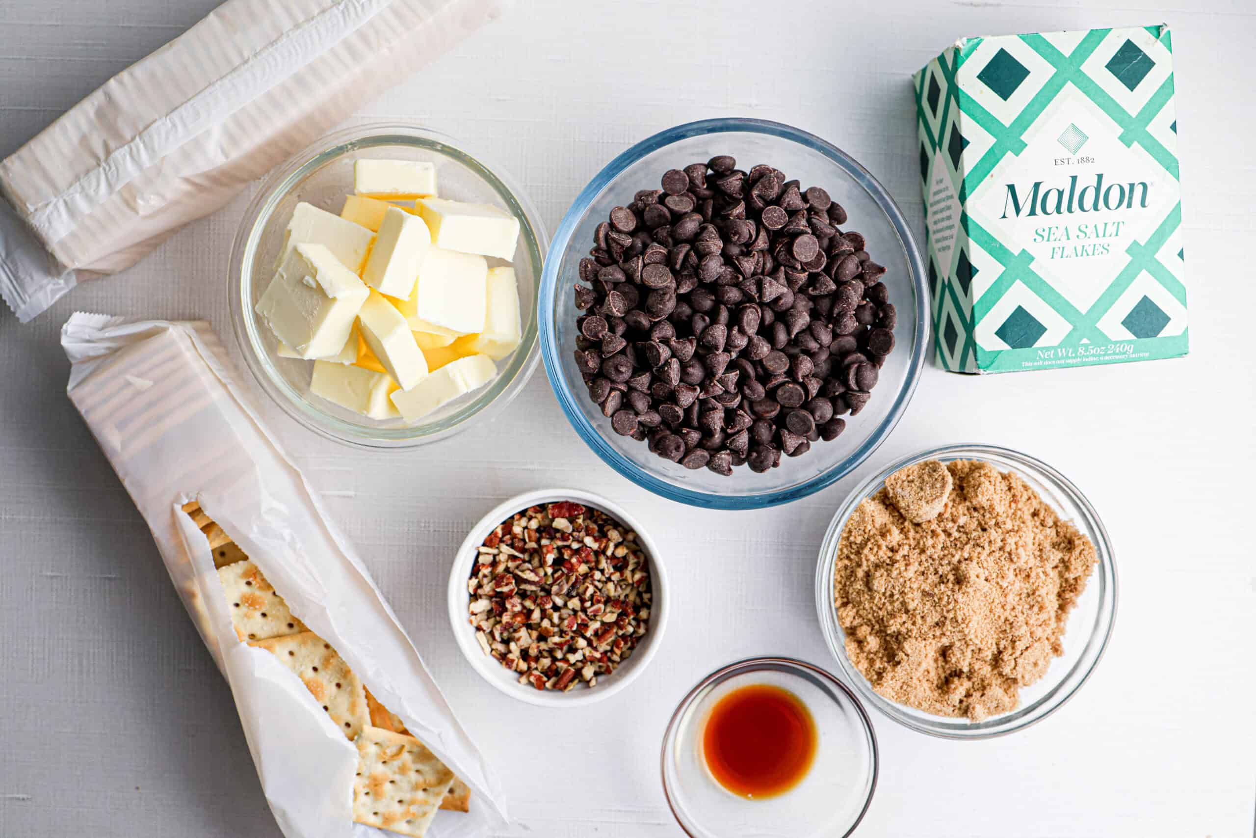 Various baking ingredients for Saltine Cracker Toffee, including chocolate chips, butter, brown sugar, vanilla extract, chopped pecans, salt, and saltine crackers arranged on a white surface.