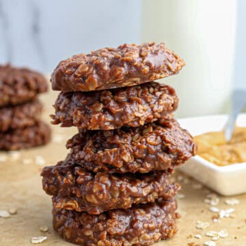 A stack of five chocolate oatmeal No Bake Cookies sits on parchment paper, with oats scattered around and a dish of peanut butter in the background.
