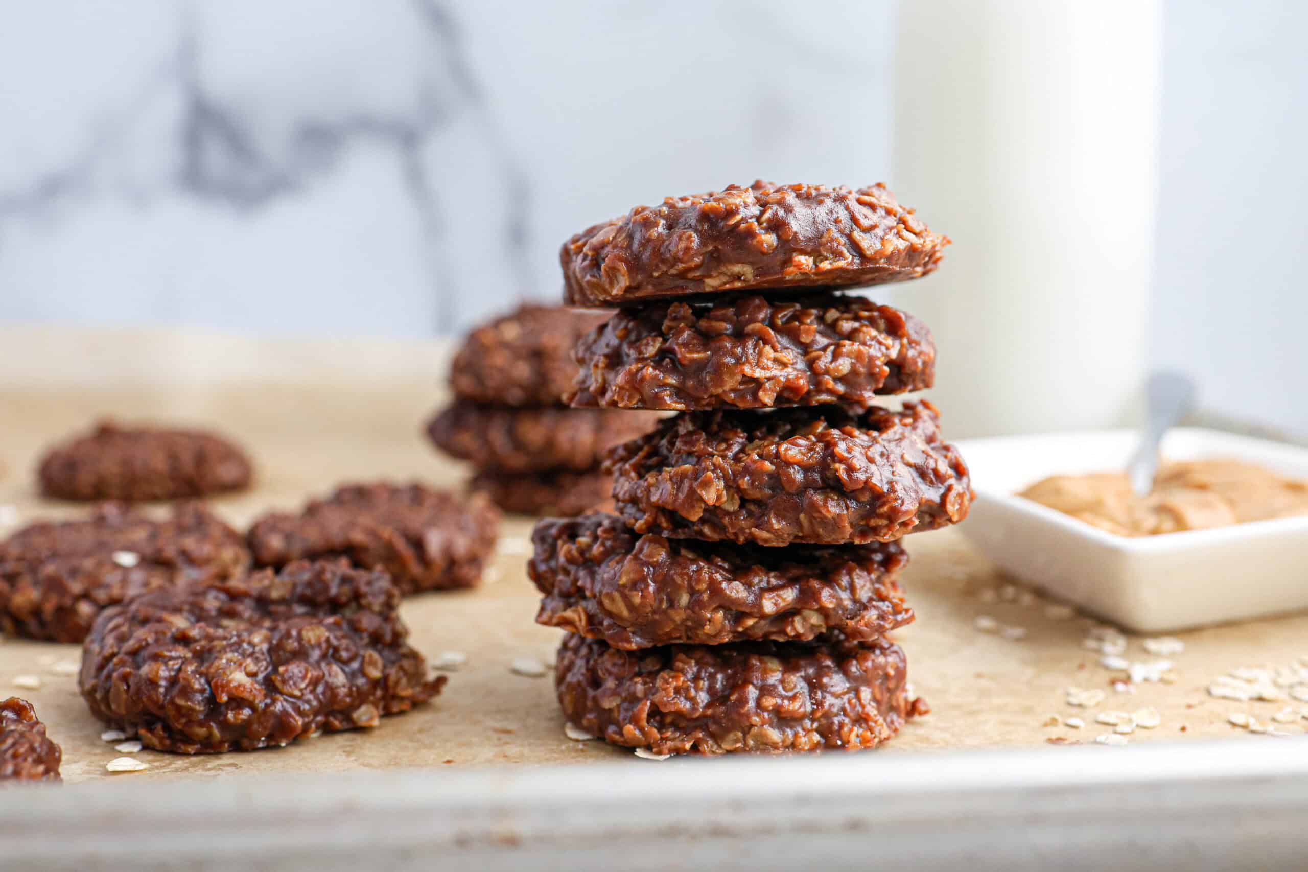 A stack of classic homemade No Bake Christmas Cookies sits on parchment paper, with more cookies, a dish of peanut butter, and a glass of milk in the background.