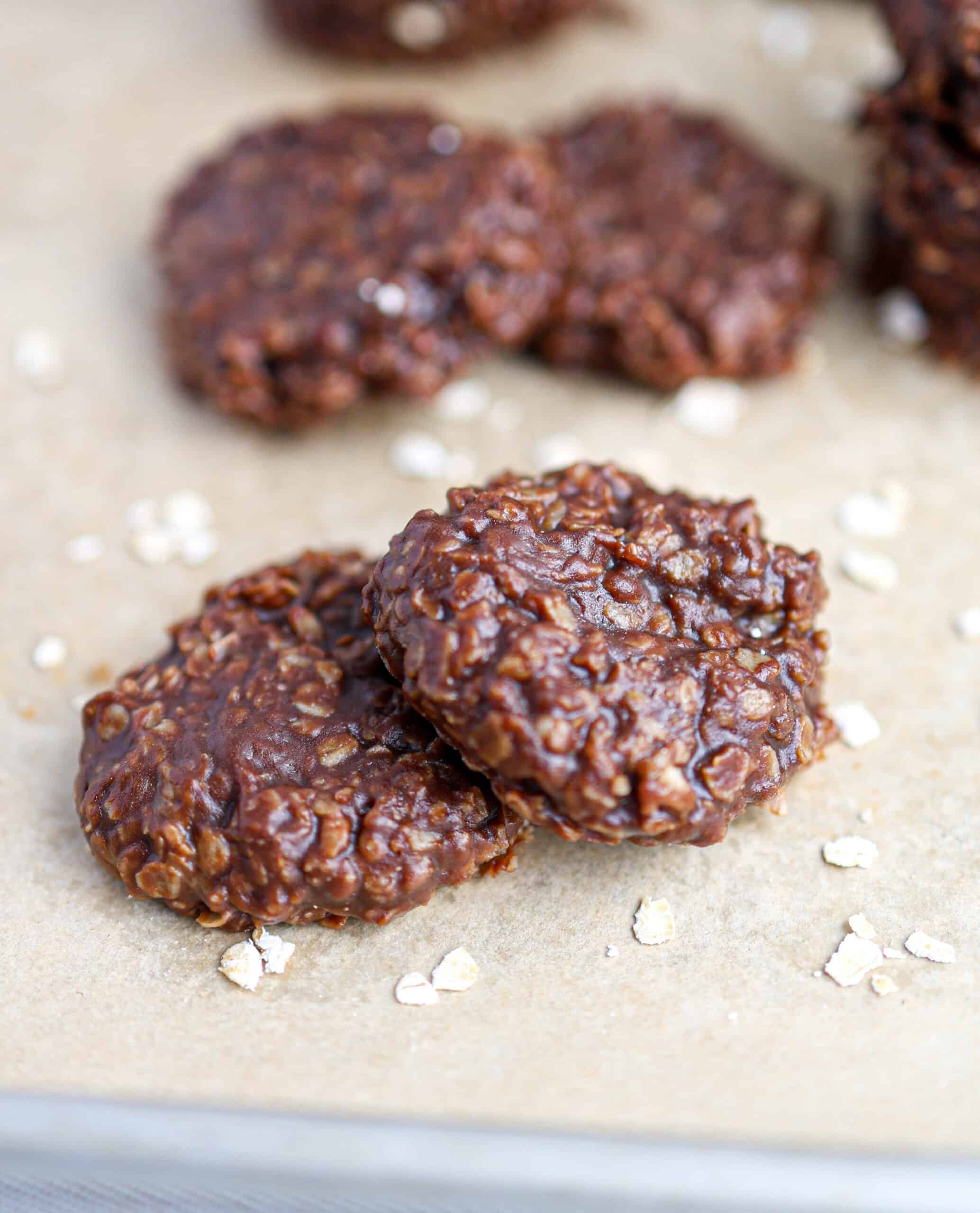 Two No Bake Cookies rest on parchment paper, their chocolate oatmeal goodness on display, with more cookies and scattered oats in the background.