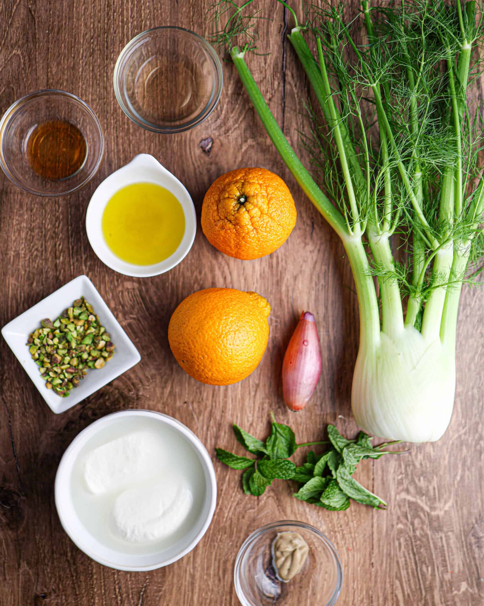 A flat lay of ingredients for a citrus and fennel salad with burrata—featuring oranges, shallot, mint, pistachios, yogurt, olive oil, mustard, vinegar, and seasonings—arranged on a wooden surface.