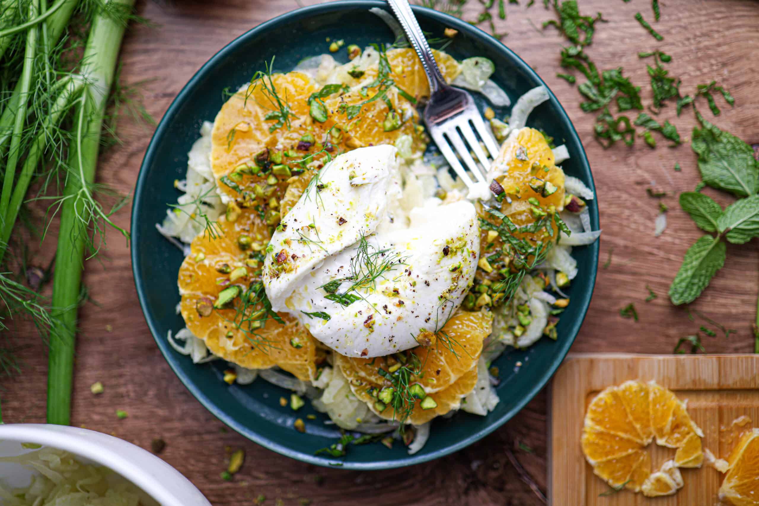 A vibrant citrus fennel salad with burrata, featuring sliced oranges, fennel, and pistachios topped with creamy burrata cheese and fresh herbs, served with a fork on the side.