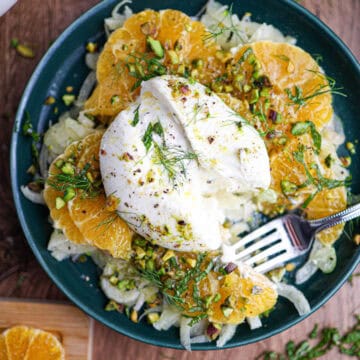 A plate of citrus and fennel salad with burrata, topped with chopped pistachios and fresh herbs, features sliced oranges and a creamy ball of burrata cheese, with a fork resting on the side.