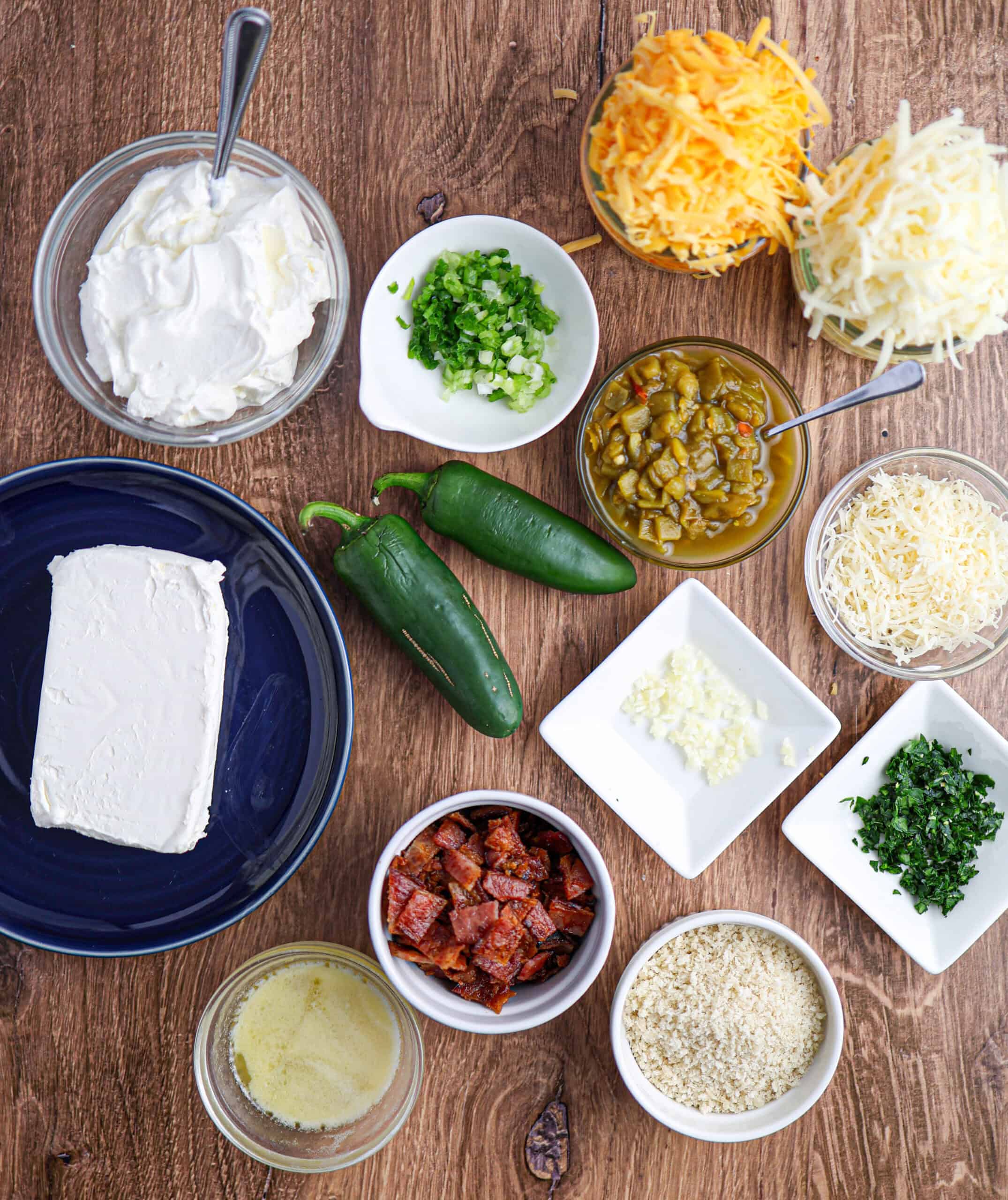 Assorted ingredients on a wooden surface, including cream cheese, shredded cheeses, green onions, jalapeños, chopped herbs, diced sausage, breadcrumbs, and minced garlic—perfect for making Jalapeño Popper Dip.