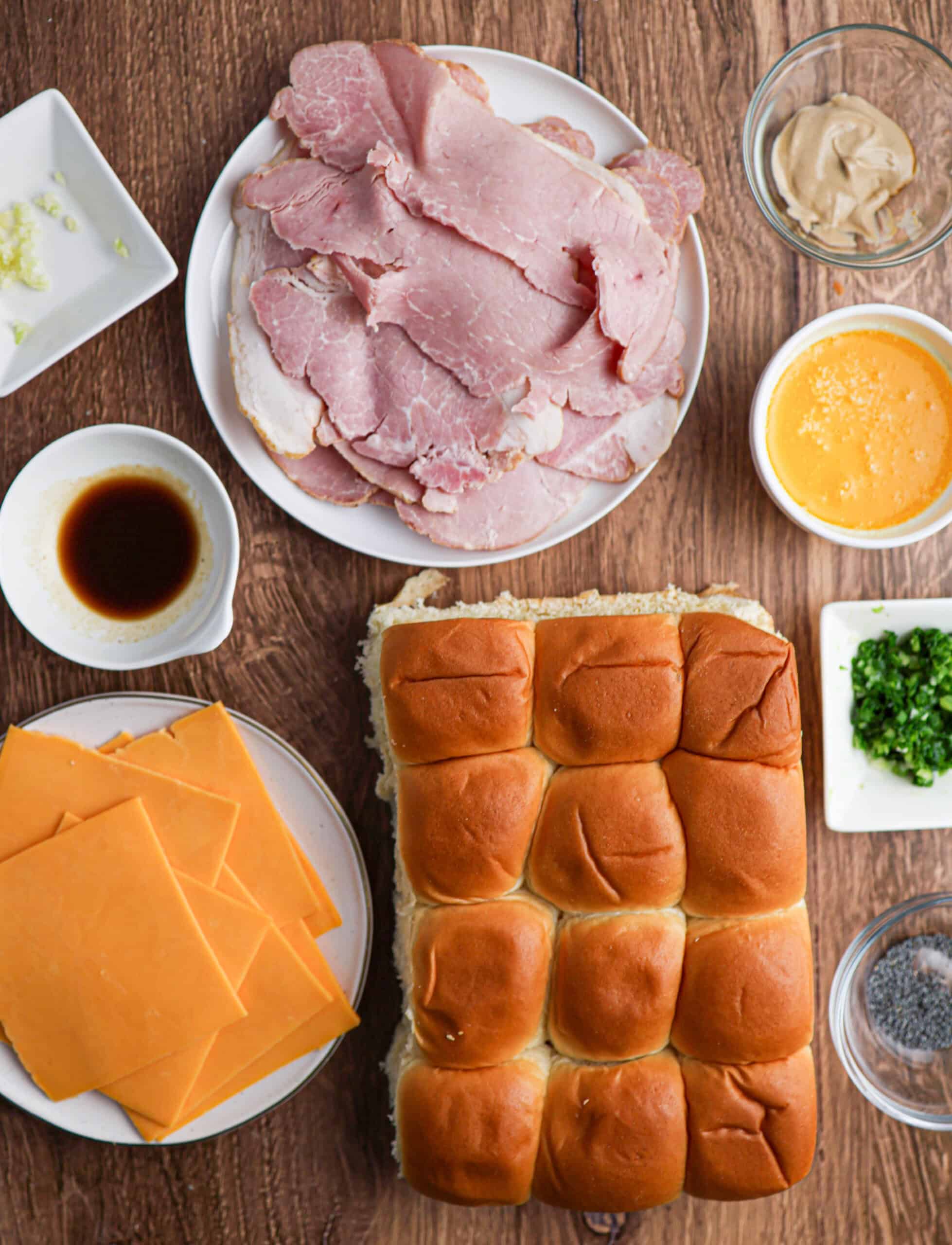 Ingredients for ham and cheese sliders are arranged on a wooden surface, featuring ham, cheddar cheese slices, slider buns, butter, mustard, Worcestershire sauce, garlic, green onions, and poppy seeds.